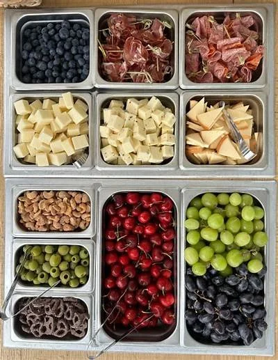 A metal tray with multiple sections filled with various fresh fruits and cheeses. Top row has blueberries, prosciutto, and ham. Middle row has cubed cheeses, sliced cheese, and a cheese wedge. Bottom row has walnuts, red cherries, green grapes, black grapes, and black olives.
