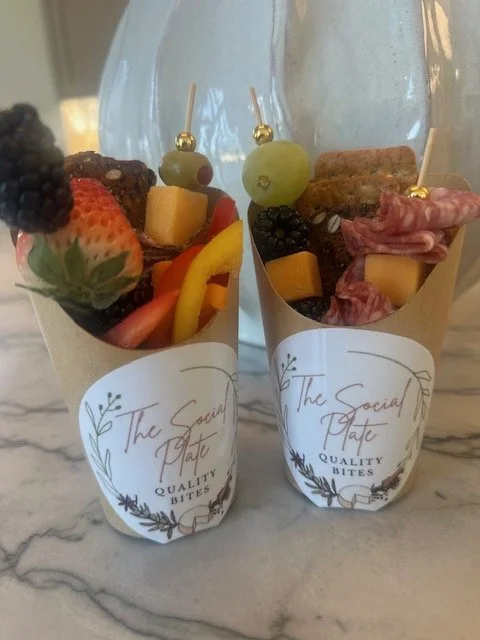 Two cones of mixed fruit, cheese, and charcuterie, wrapped with paper labeled "The Social Bite Quality Bites," placed on a marble surface with a glass pitcher in the background.
