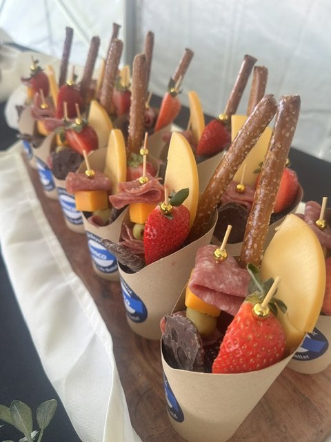 Individual dessert cups with strawberries, cheese slices, chocolate pieces, pretzel sticks, ham, and a small leaf, placed on a wooden serving board.