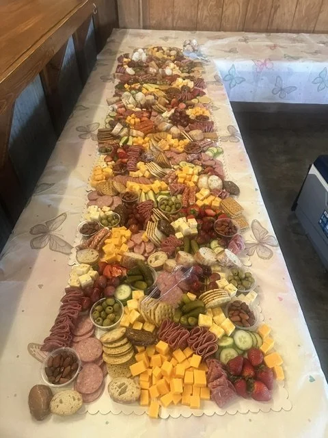 A long table covered with a butterfly-patterned tablecloth, displaying an assortment of cheeses, meats, fruits, and snacks arranged for a party or event.