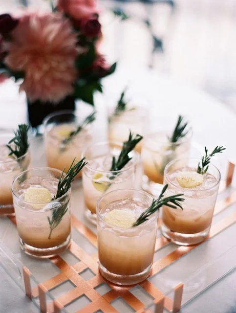 Six glasses of a creamy drink garnished with lemon slices and sprigs of rosemary on a copper tray, with a blurred flower arrangement in the background.