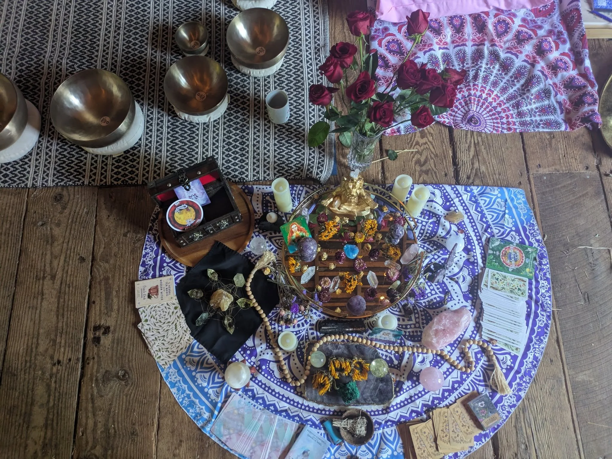 An arrangement of spiritual and mystical objects on a cloth-covered table, including roses, candles, crystals, tarot cards, and a small hand drum, possibly for a ritual or ritualistic display.