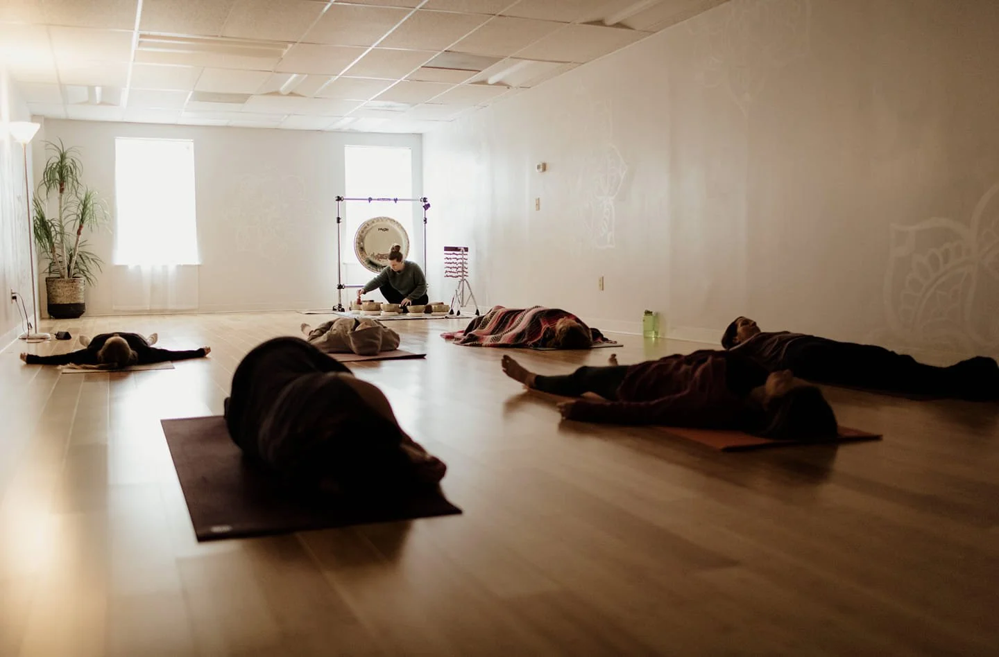 People practicing yoga in a dimly lit room with large windows, mats, and a yoga instructor with a gong in the background.