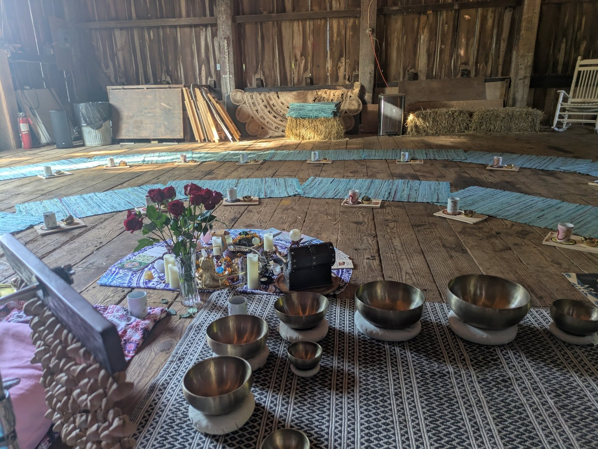 Indoor space arranged for a peaceful gathering, featuring blue patterned mats, candles, and ritual bowls on a wooden floor, with a bouquet of red roses and various decorative items at the center.