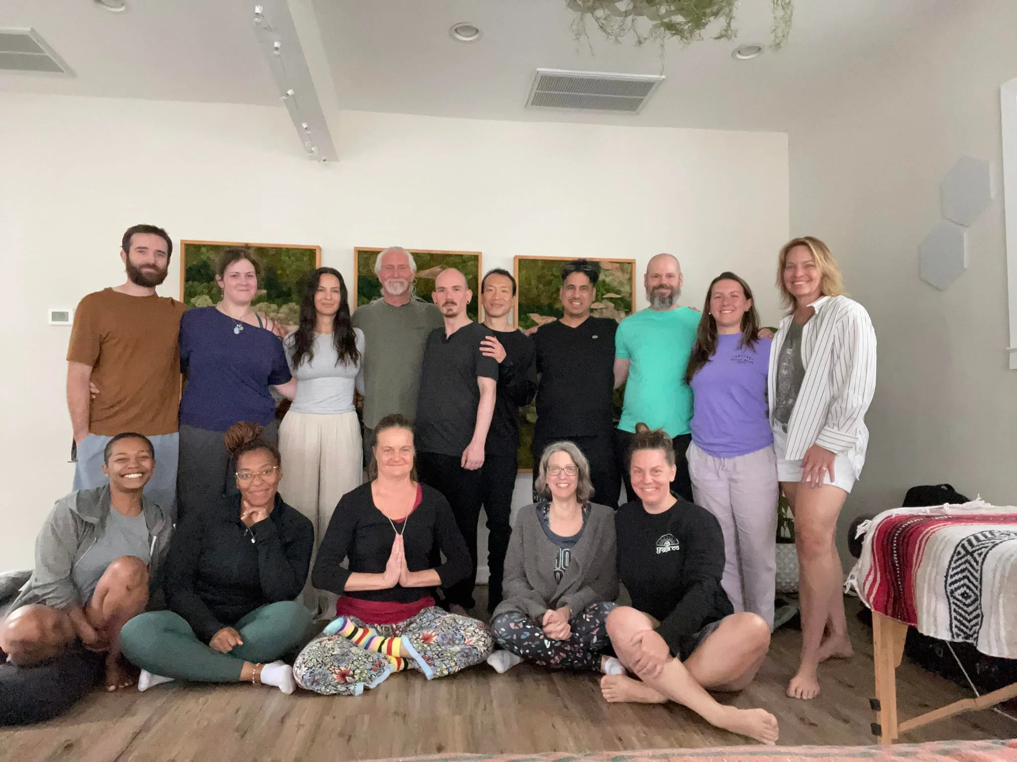 A group of fifteen people, including men and women, posing together indoors in front of a white wall with landscape paintings. Some are standing, some are kneeling on the floor, and one woman on the right is standing barefoot next to a table with a patterned cloth.