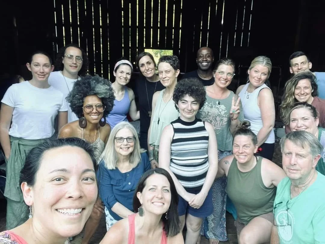 Group of 15 diverse people smiling indoors, some making peace signs, with a rustic wooden wall in the background.