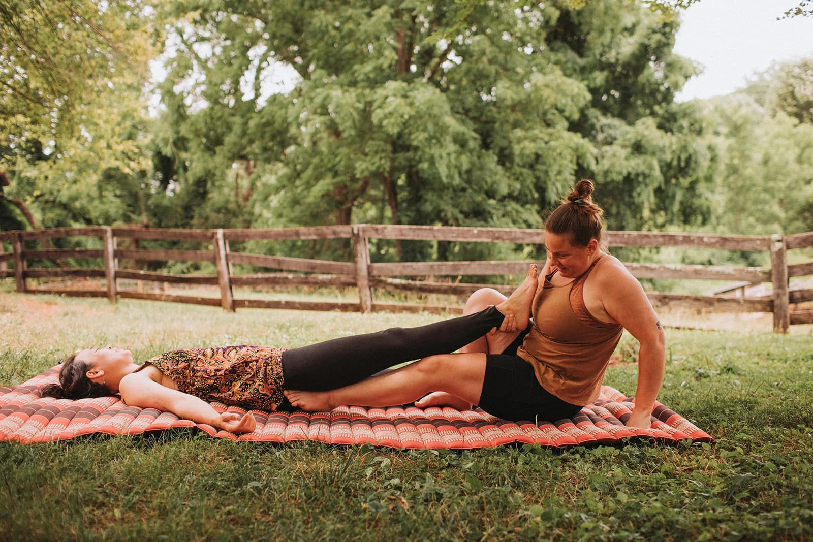 Two women practicing acro yoga outdoors on a mat on grass, with one woman lying on her back and the other woman sitting on her partner's lap, supporting her by holding her foot, amidst a natural setting with trees and a wooden fence.