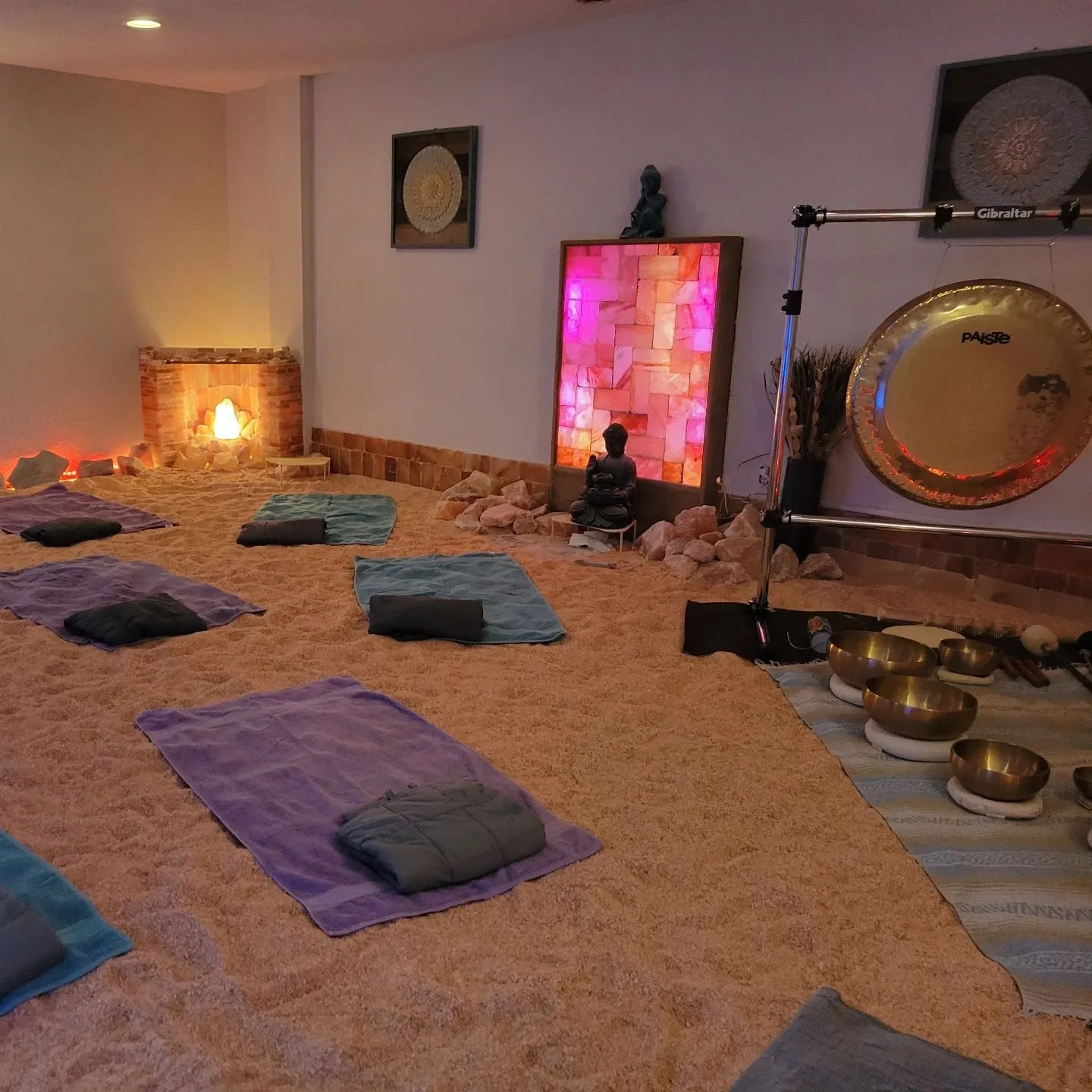 A sacred space with yoga mats and meditation cushions on a warm orange carpet, illuminated by soft, ambient lighting, decorated with statues, a glowing salt lamp, lighted wall art, a large gong, singing bowls, and spiritual artwork.