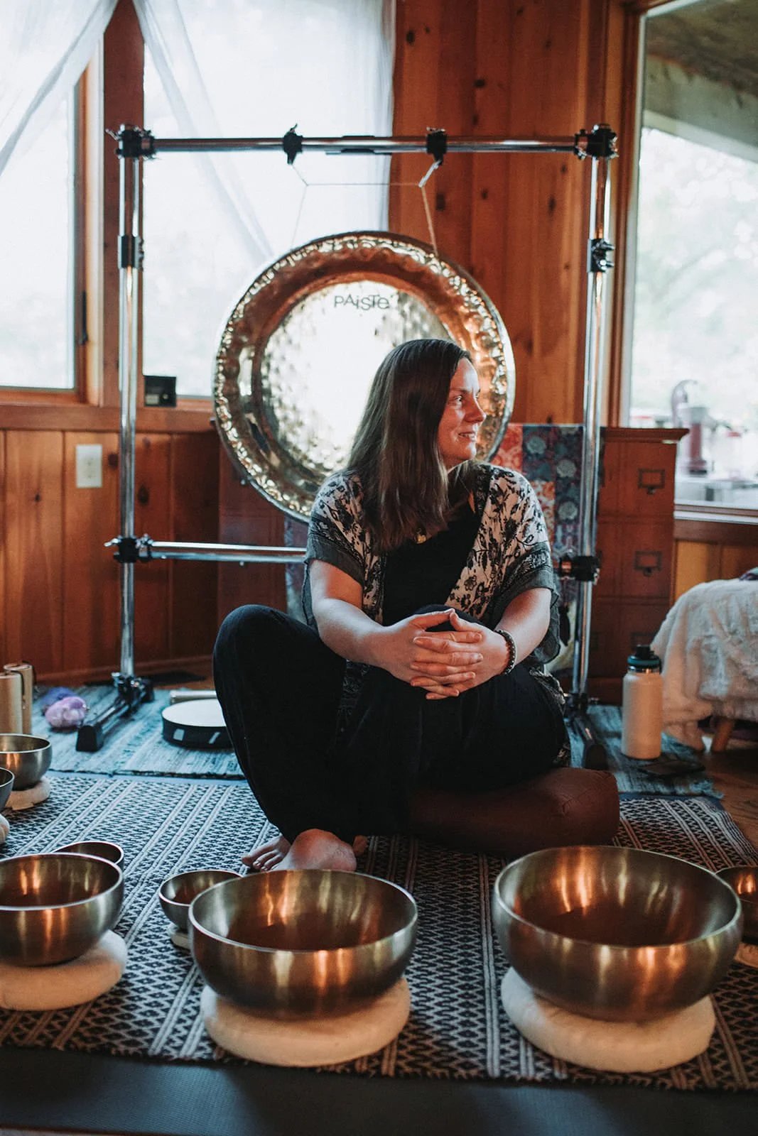 A woman sitting cross-legged on a cushion on the floor, surrounded by singing bowls, indoors with wooden walls and large windows, with a large metal gong behind her.