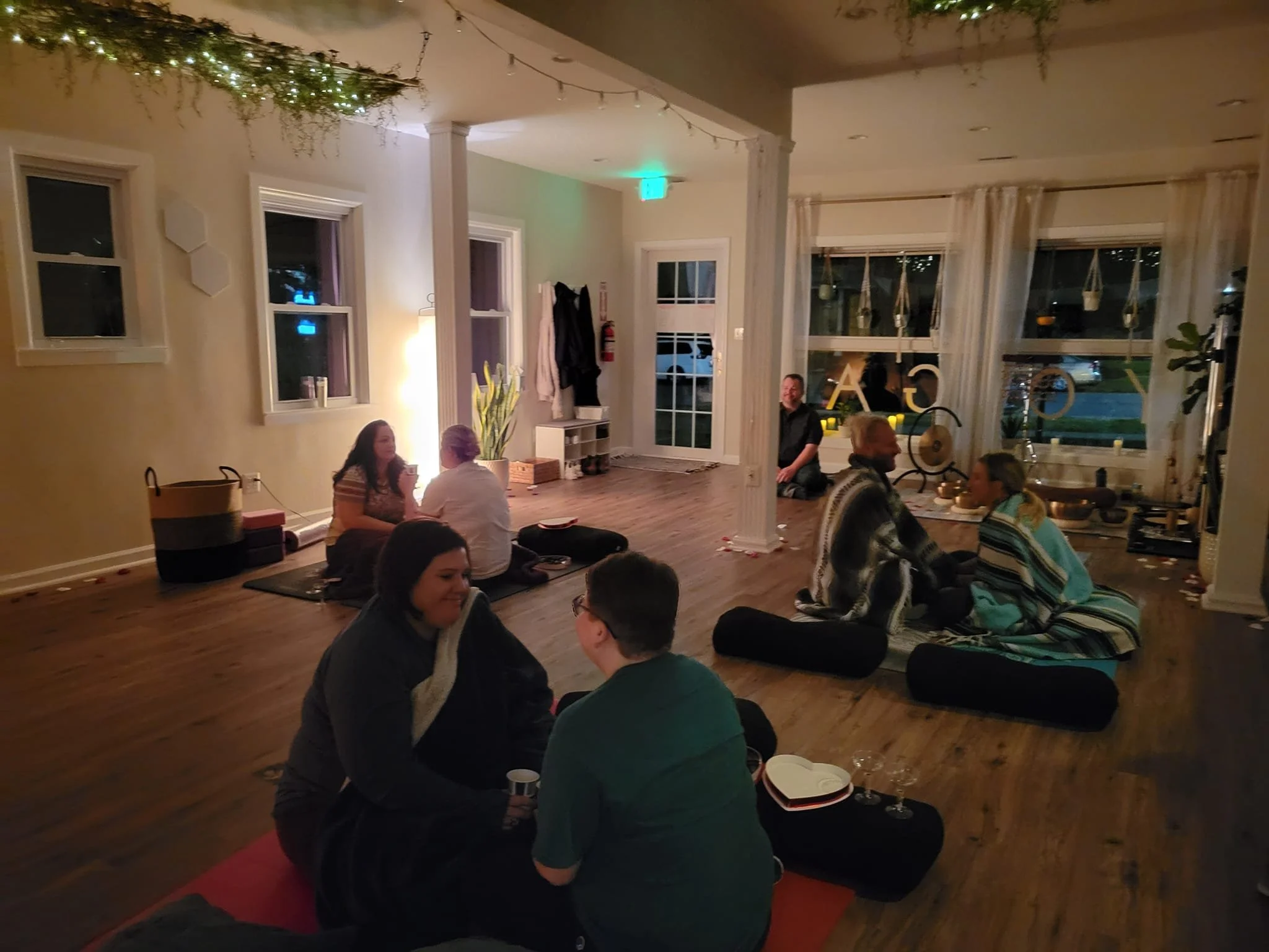A group of people gathered in a cozy, warmly lit living room, sitting on the floor with cushions, decorated with string lights and hanging plants, enjoying social time.