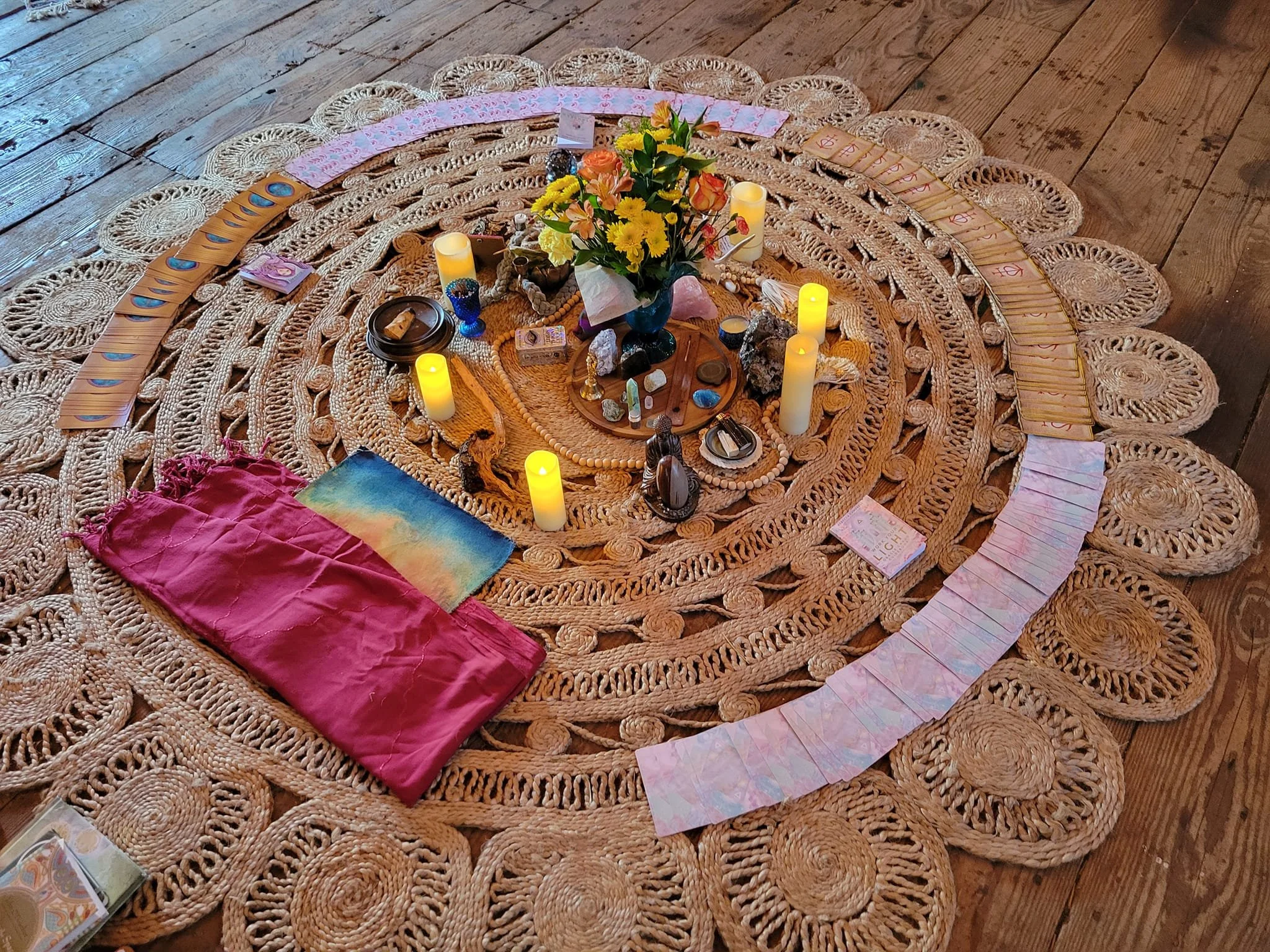 A circular woven rug with spiritual and tarot items arranged in the center. The setup includes a bouquet of yellow and orange flowers, candles, crystals, tarot cards, and small statues, creating an altar or sacred space.