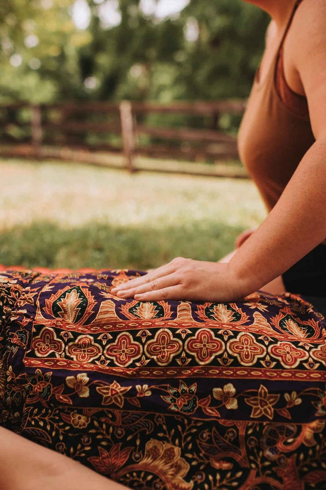 Person practicing yoga outdoors on a colorful patterned blanket, with green trees and a wooden fence in the background.