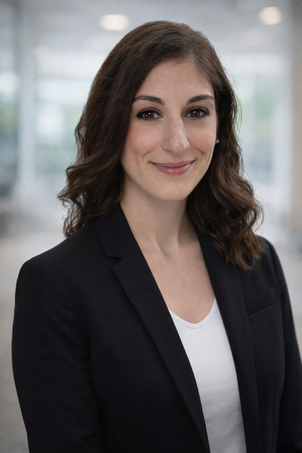 A professional woman with shoulder-length brown hair and brown eyes, wearing a black blazer over a white top, smiling at the camera in an indoor setting.