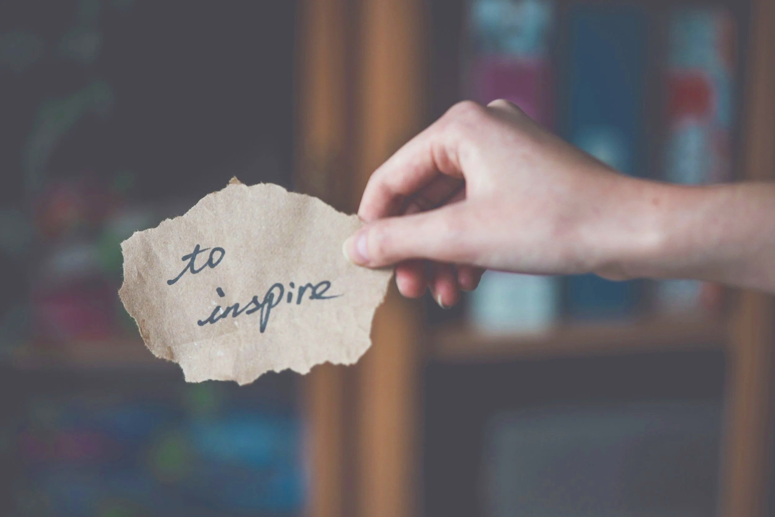 A hand holding a torn piece of paper with the words 'to inspire' written on it, against a blurred background of bookshelves.