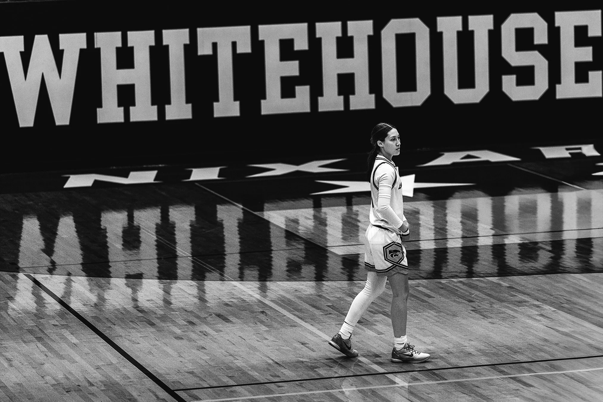 A woman wearing athletic clothing walking on a basketball court with a large ‘White House’ sign in the background, reflected on the polished floor.
