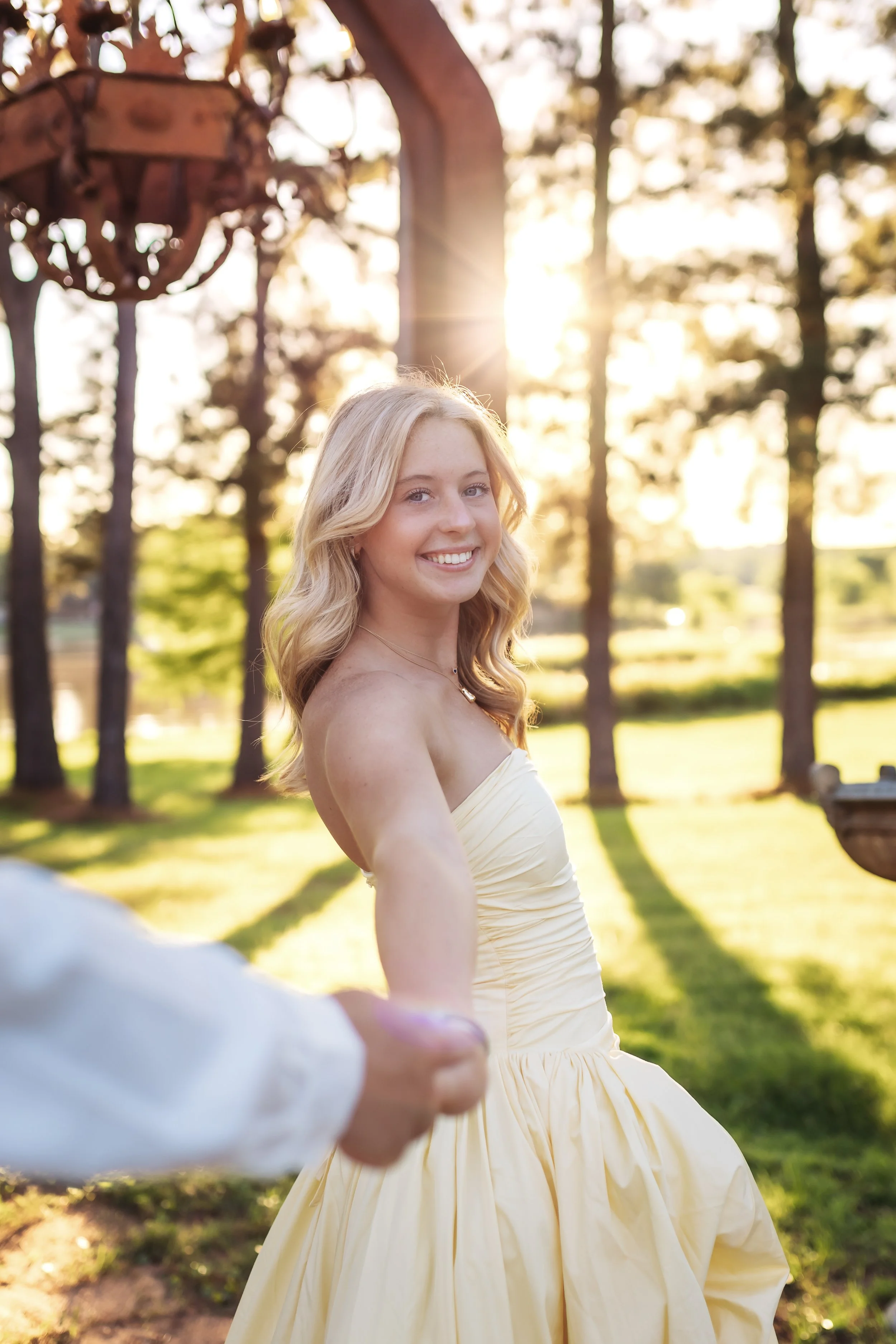 A blonde woman in a strapless yellow dress holding hands with someone outside in a sunlit park with trees in the background.