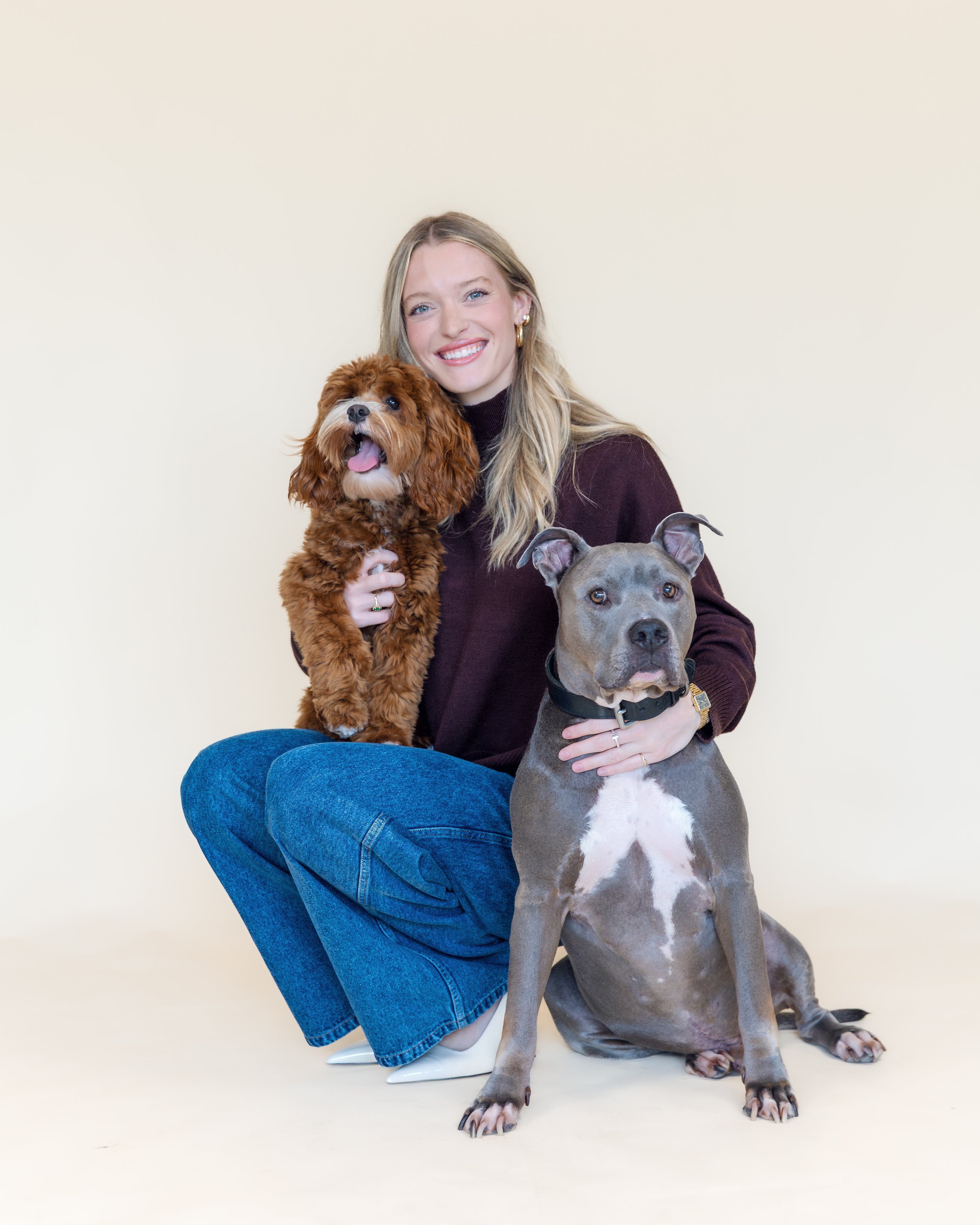 A woman sitting on the floor with two dogs, one sitting on her lap and the other sitting on the ground, against a plain light-colored background.