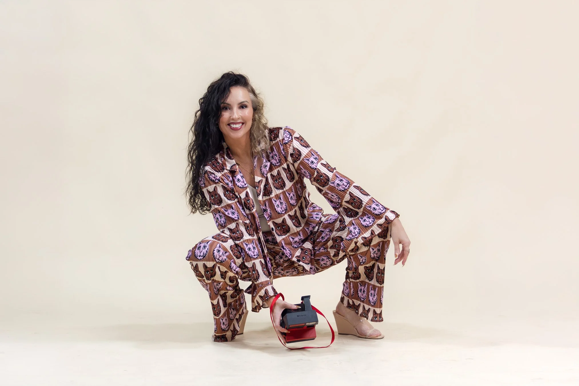 Woman in patterned pajamas squatting and holding a vintage camera, smiling against a plain background.