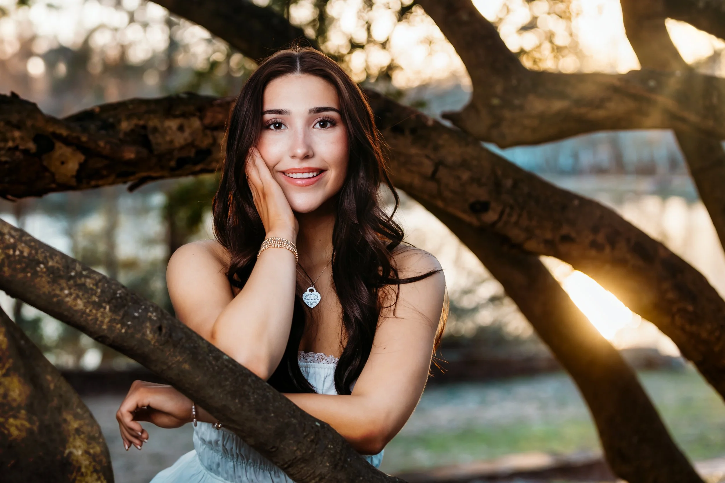 Young woman with long dark hair, smiling, posing outdoors among tree branches during sunset, wearing a white dress and jewelry.