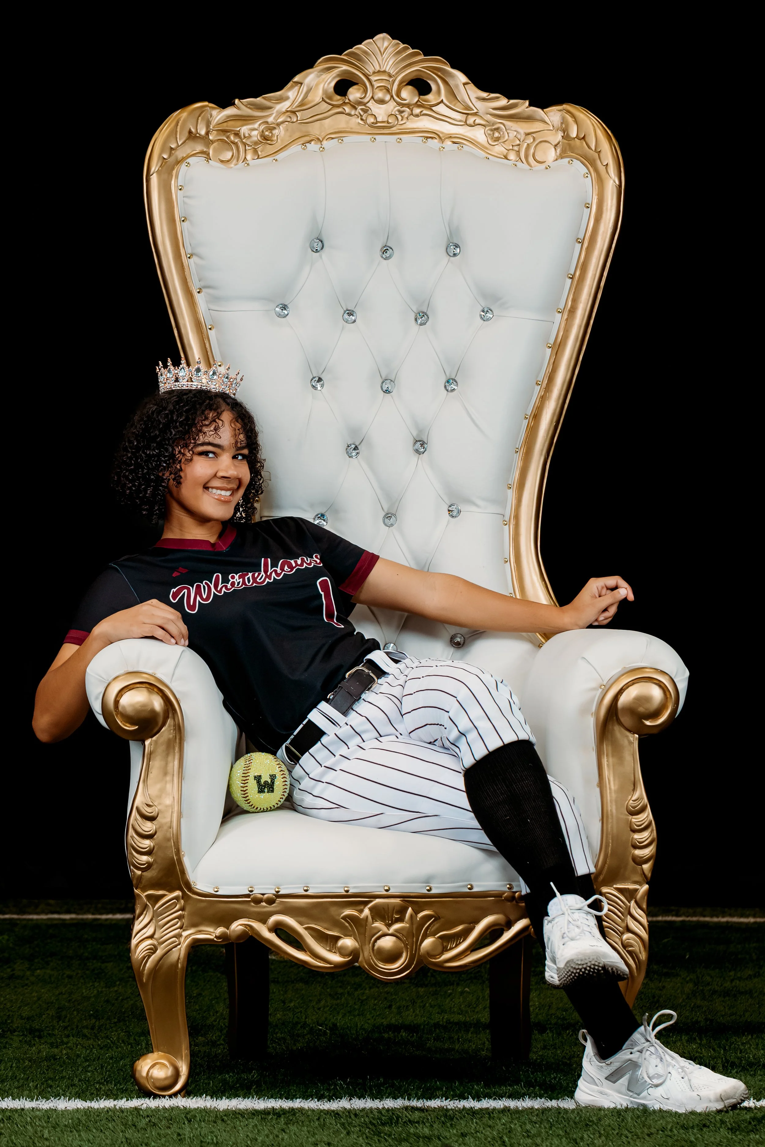 Young woman with curly hair, wearing a black baseball uniform with 'Whitehouse' on it, sitting on a large ornate white and gold throne-like chair, wearing a silver crown, with a softball and a baseball glove nearby, smiling at the camera.
