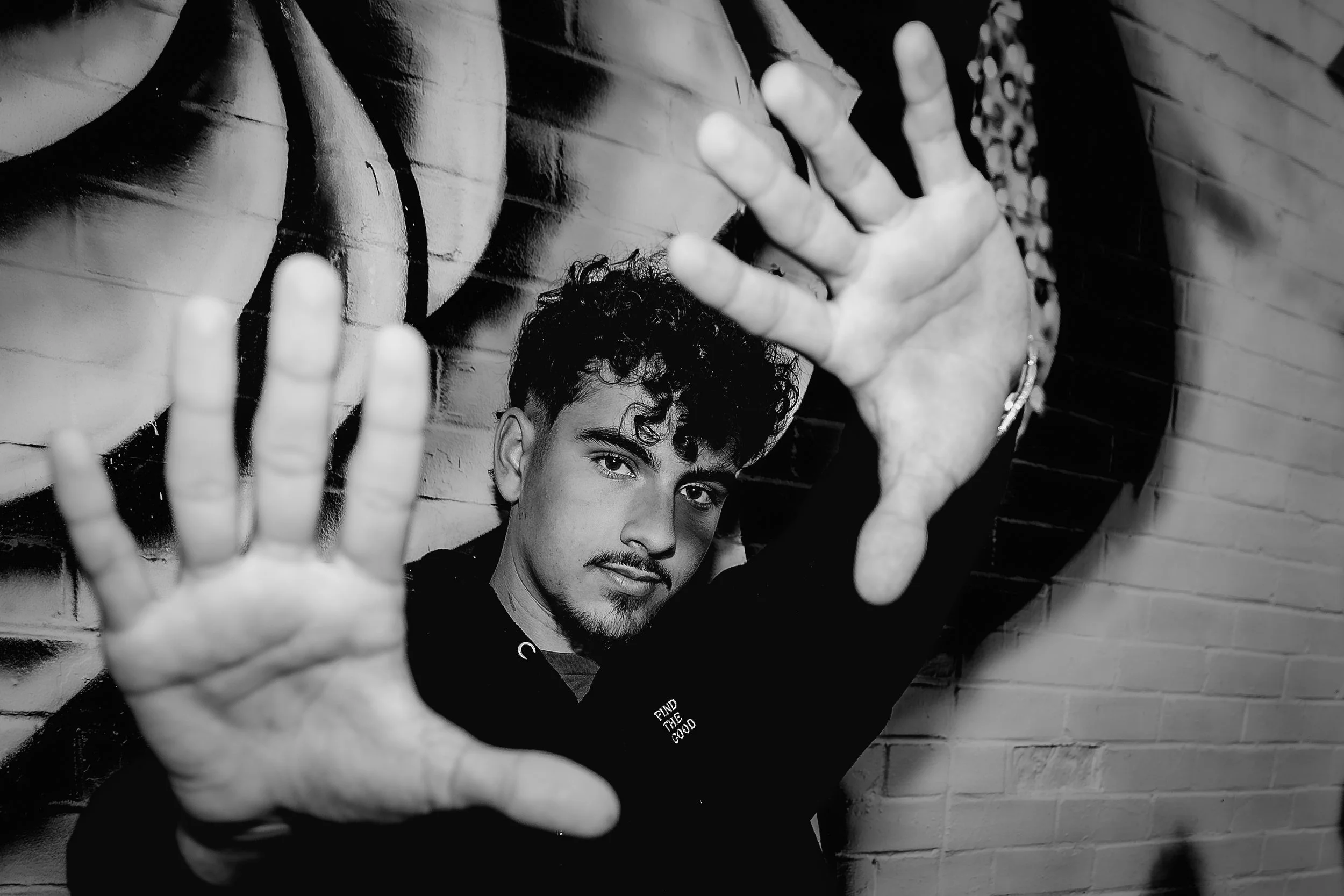 Young man with curly hair and a mustache making a frame with his hands in front of a brick wall with graffiti.