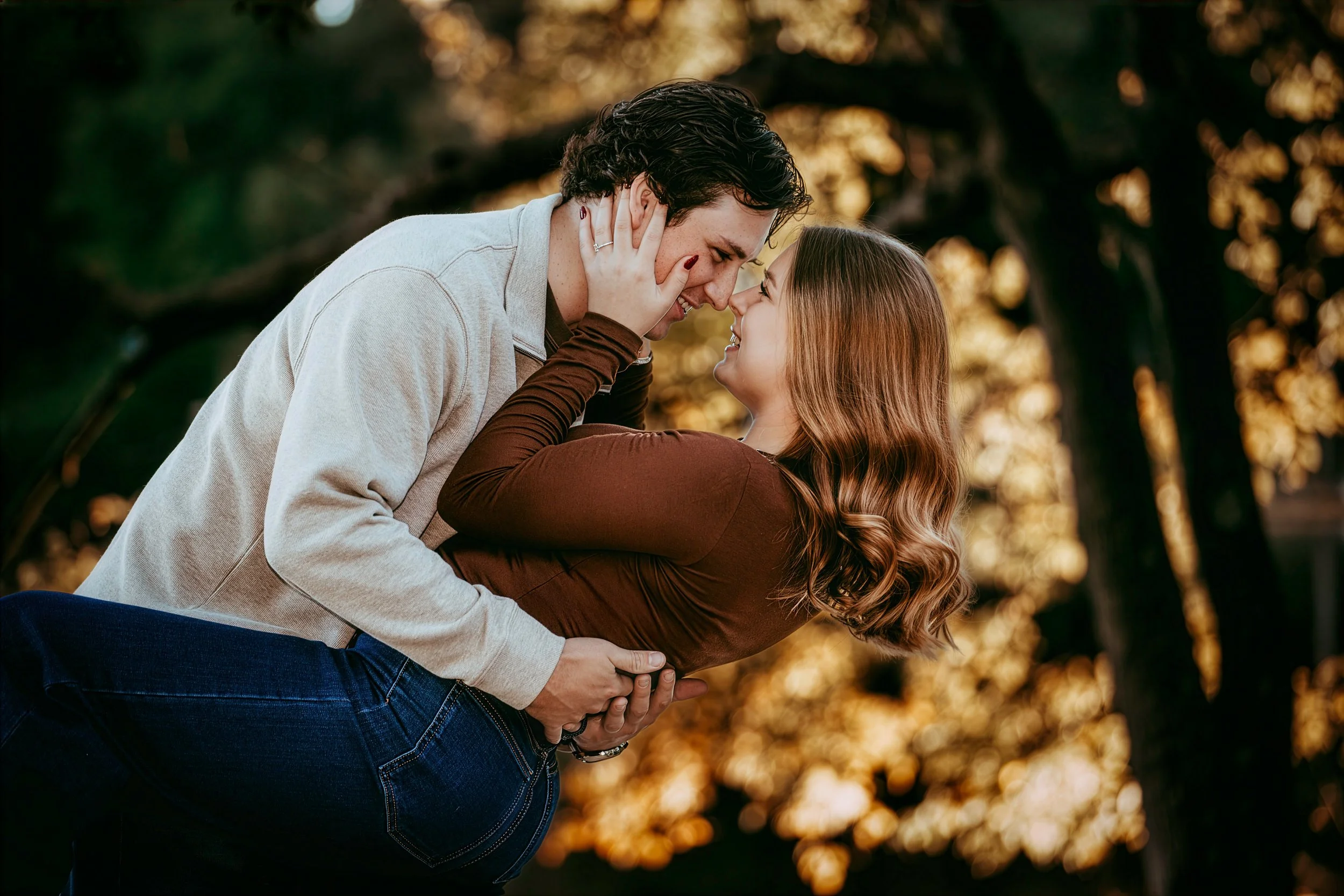 A couple embracing outdoors during sunset, with the man leaning down and touching noses with the woman, both smiling and holding each other. The background shows blurred trees with golden leaves.