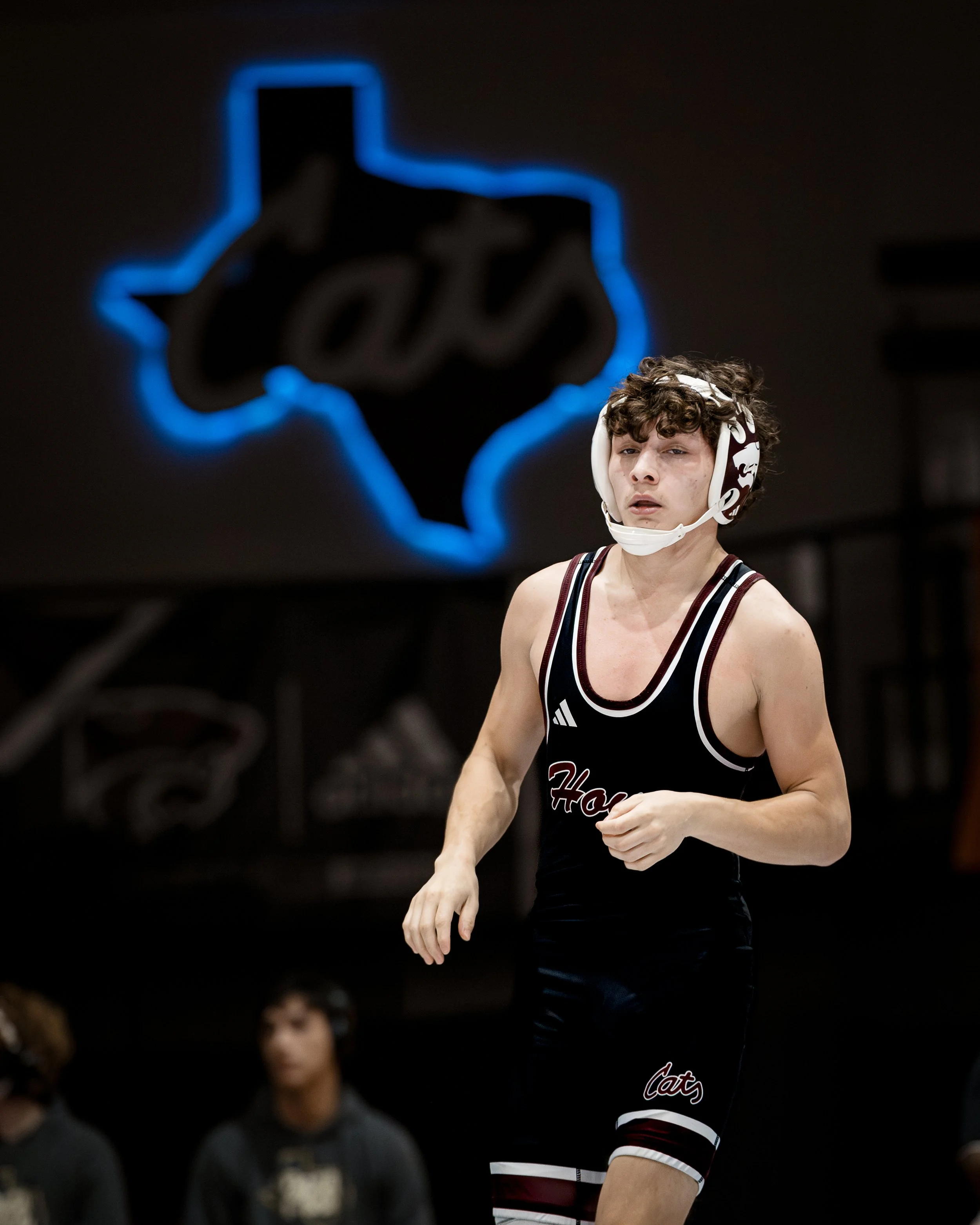 A wrestler in a black singlet with red and white accents, wearing a protective headgear and looking focused, in an indoor gym with a neon Texas outline sign in the background.