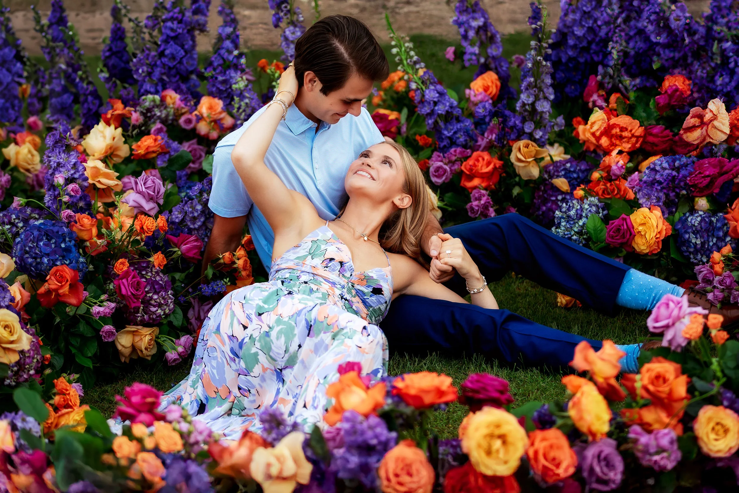 A couple sitting among colorful flowers, smiling and looking at each other affectionately.