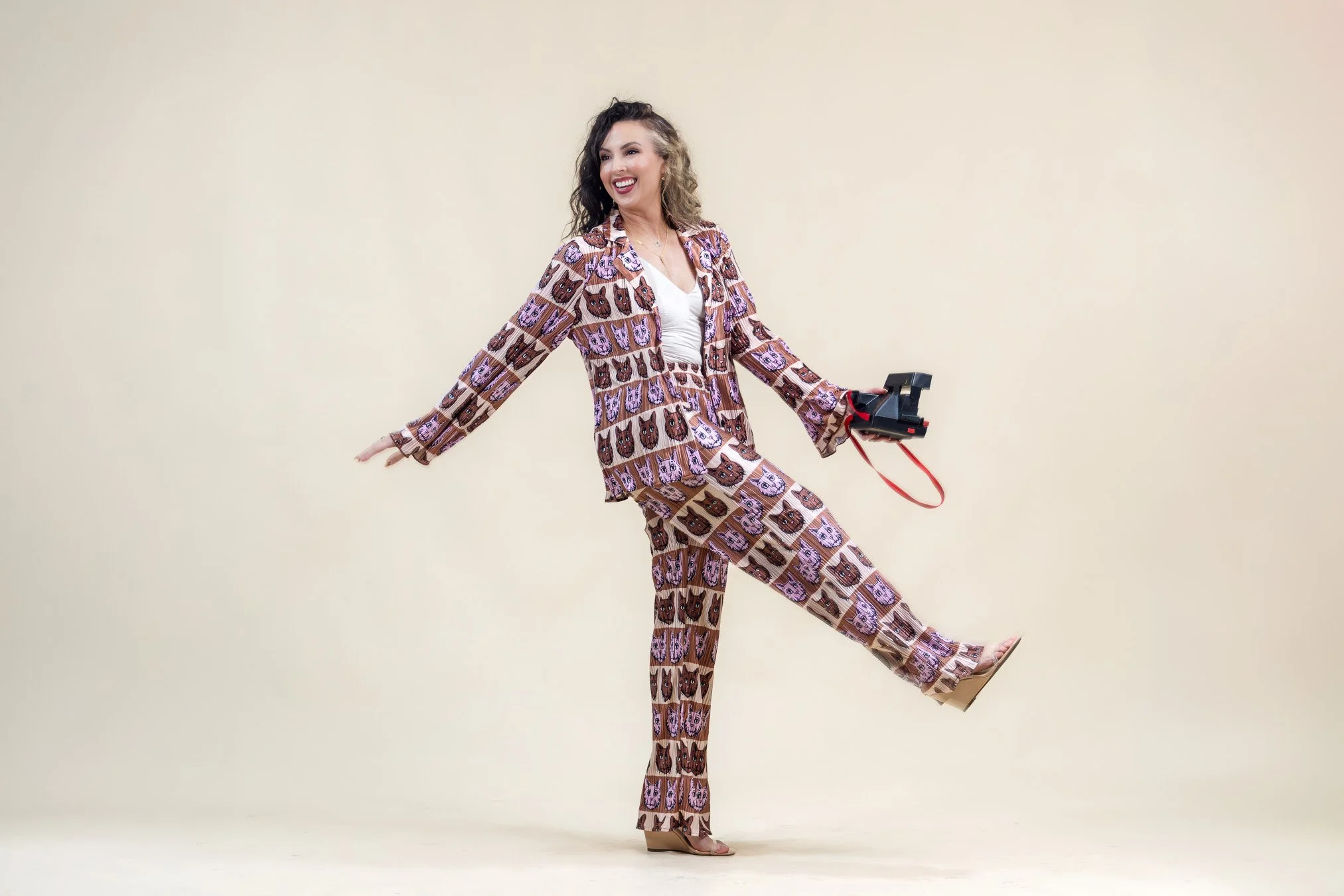 A woman smiling, wearing a matching pantsuit with a pattern of cartoon owls, holding a camera, standing against a plain beige background.