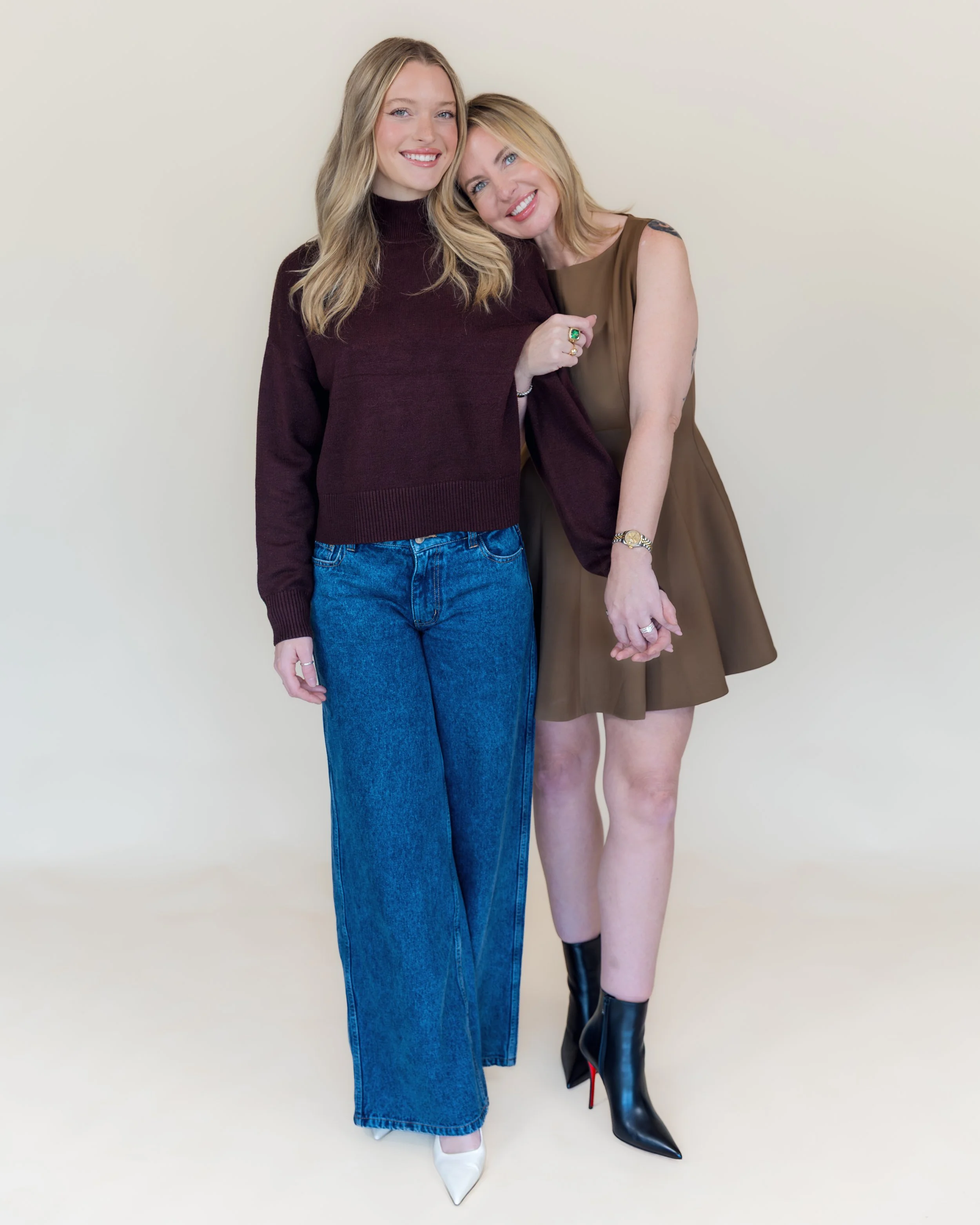 Two women standing close, holding hands, smiling, against a plain light-colored wall. One woman has long blonde hair, wears a dark turtleneck sweater and wide-legged jeans. The other has shoulder-length blonde hair, wears a sleeveless brown dress and