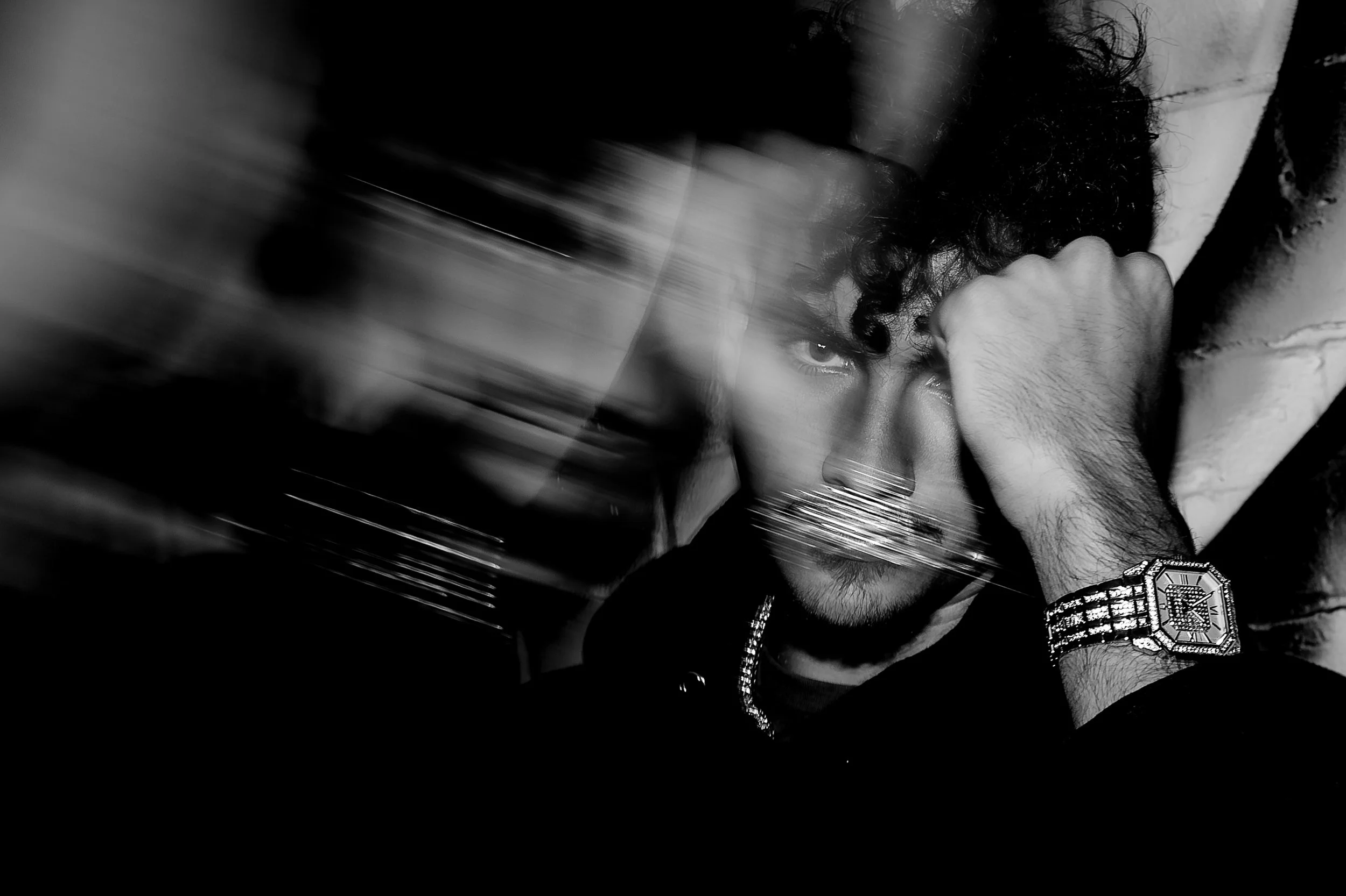 Black and white photo of a man with curly hair and a beard, wearing a watch and jewelry, resting his head on his hand with a serious expression, with a blurred motion streak across the image.