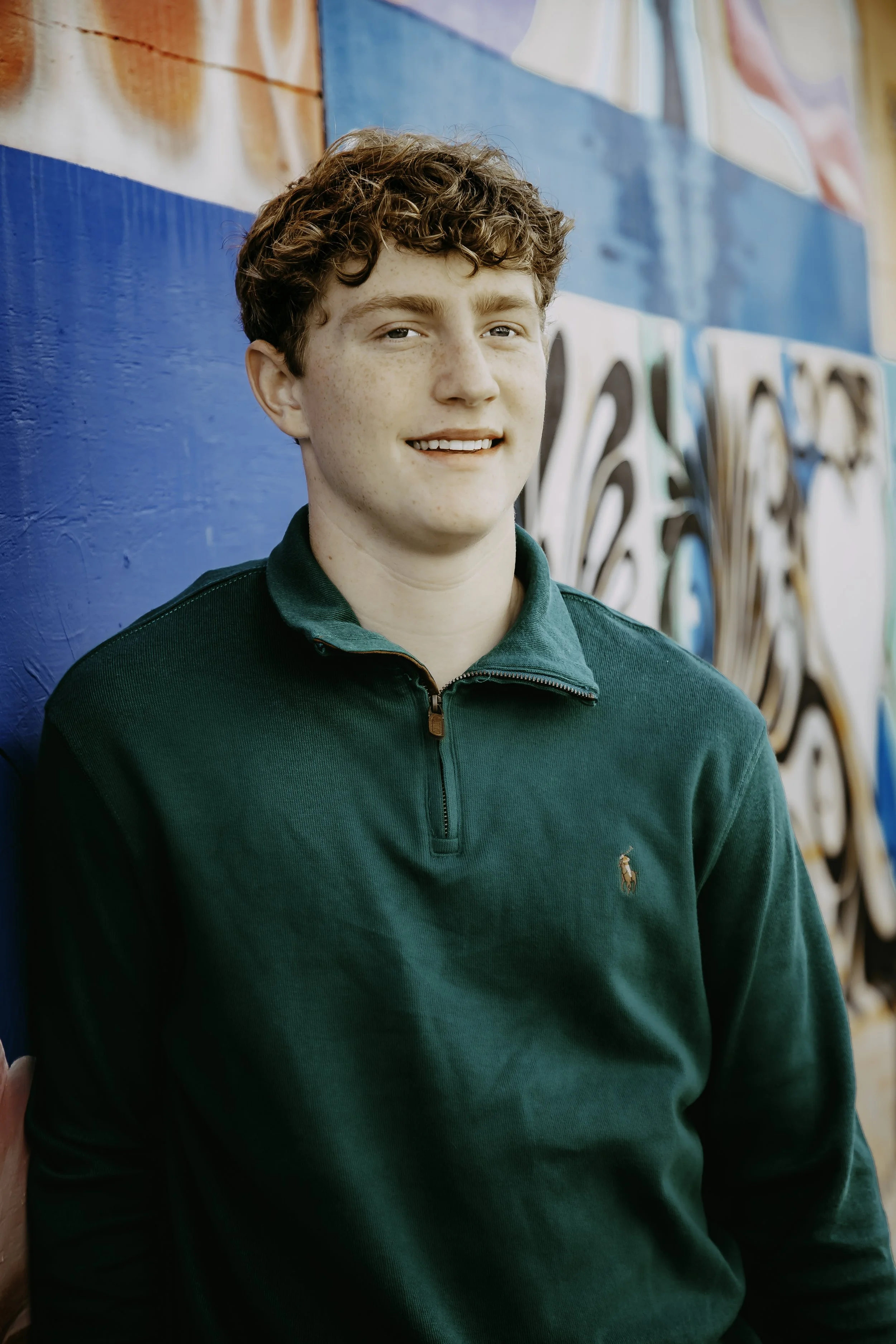 A young man with curly red hair, fair skin, and blue eyes standing against a colorful graffiti wall. He is wearing a green zip-up sweater with a small polo logo.