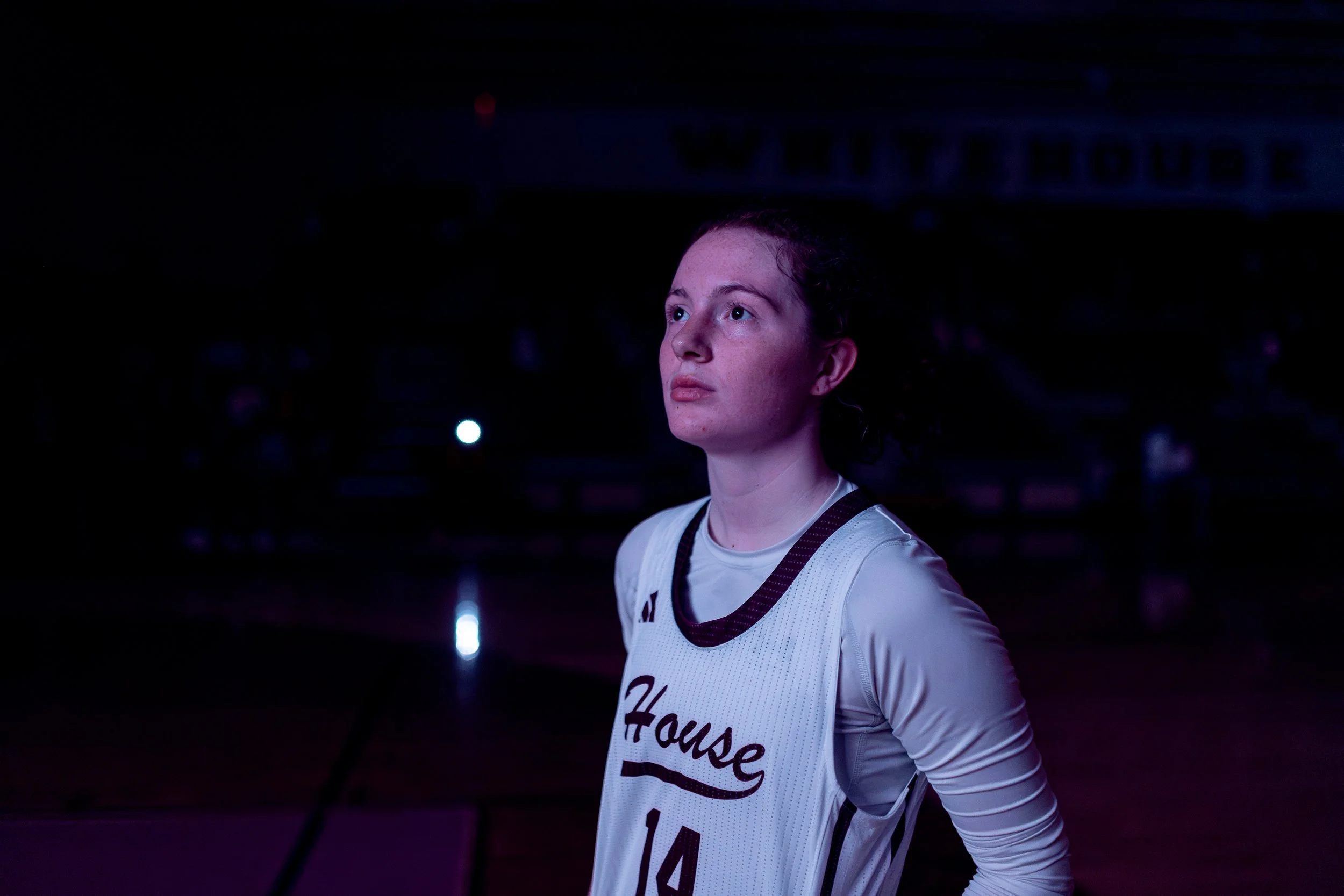 Young female basketball player with a serious expression, wearing a white jersey with the word 'House' and the number 14, standing on the court in a dark arena.