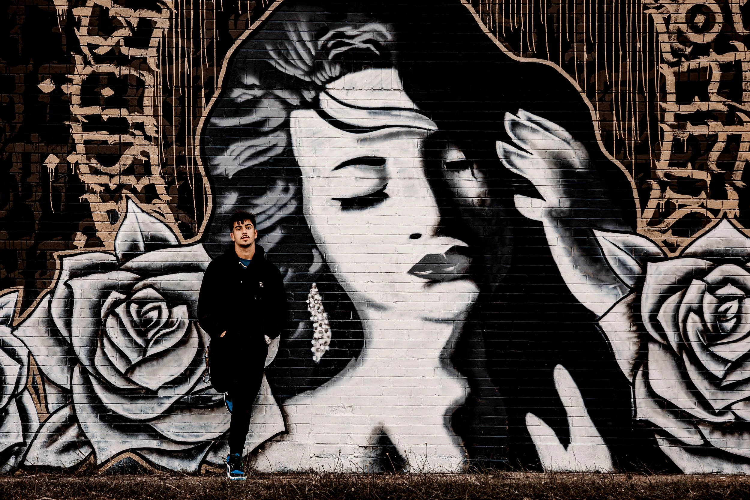 A young senior high school student standing with his hands in his jacket pockets in front of a large black and white mural of a woman with flowing hair, roses, and decorative patterns painted on a brick wall.