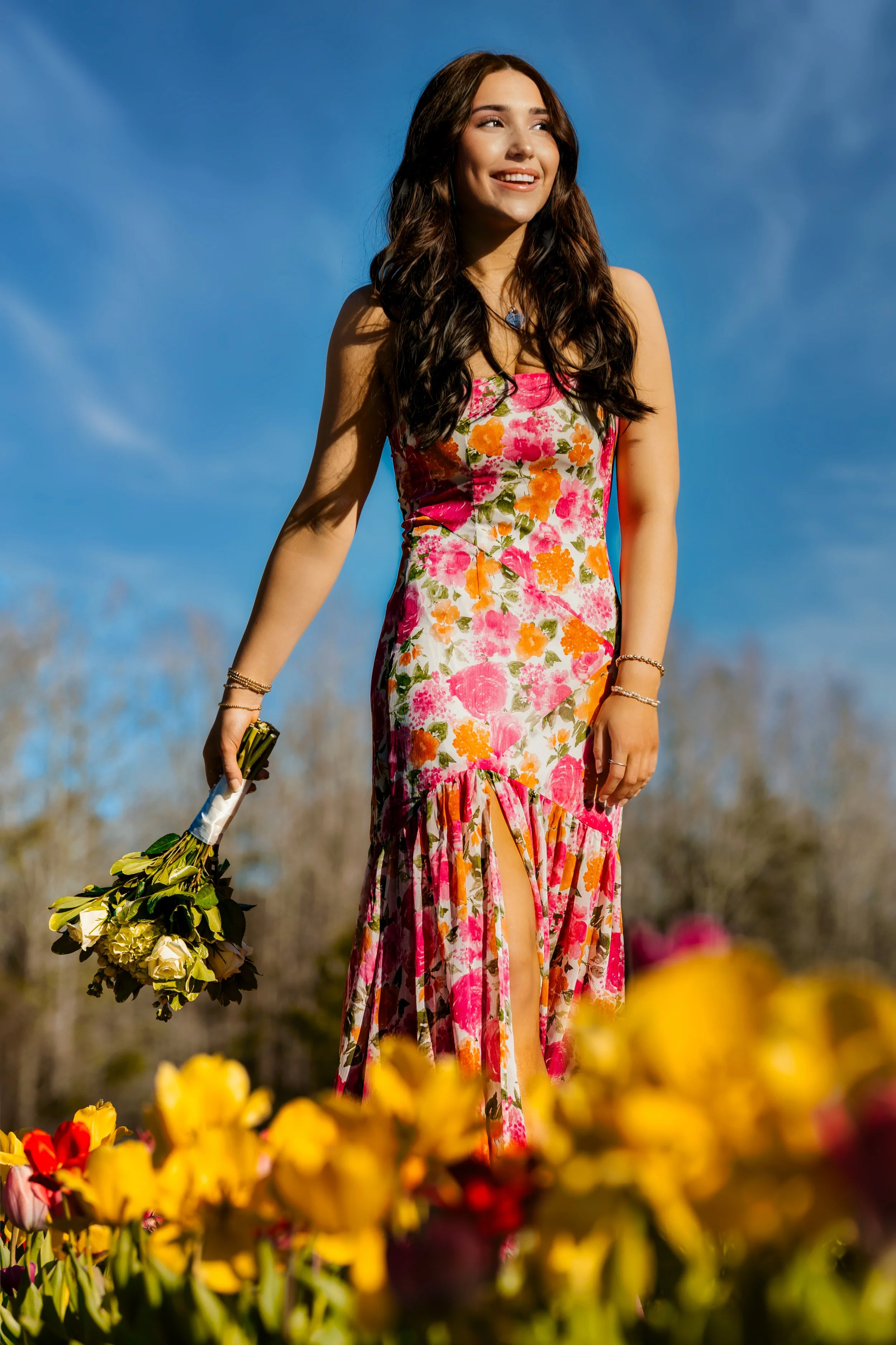A young woman with long dark hair in a floral dress holding a bouquet of flowers, standing outdoors in a flower field with a clear blue sky.