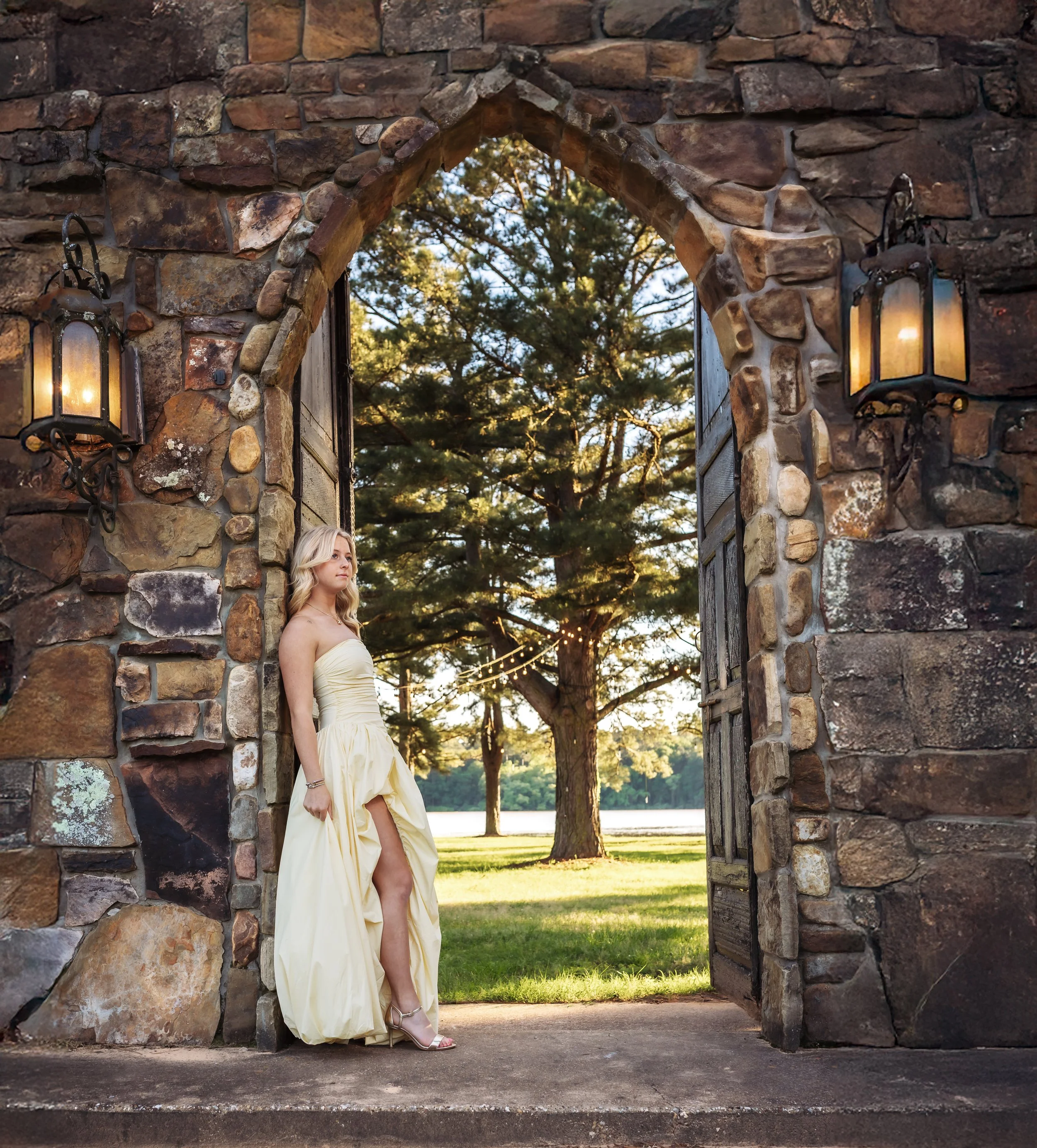 A woman in a strapless yellow dress standing in an arched stone doorway with open wooden doors, overlooking a grassy area with trees and string lights at sunset.