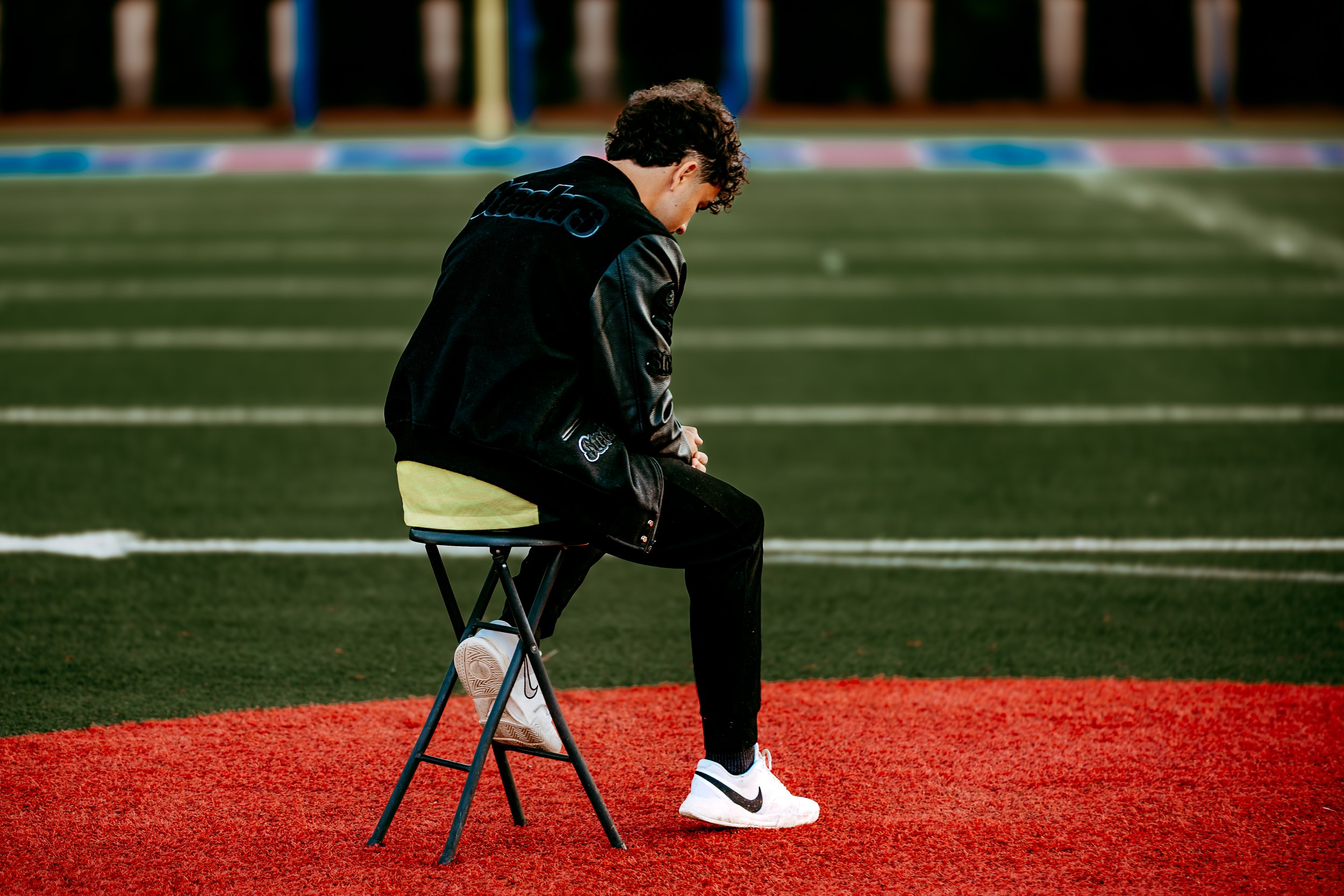 Person sitting on a chair at the edge of a sports field, looking down, wearing a black jacket and white Nike sneakers.