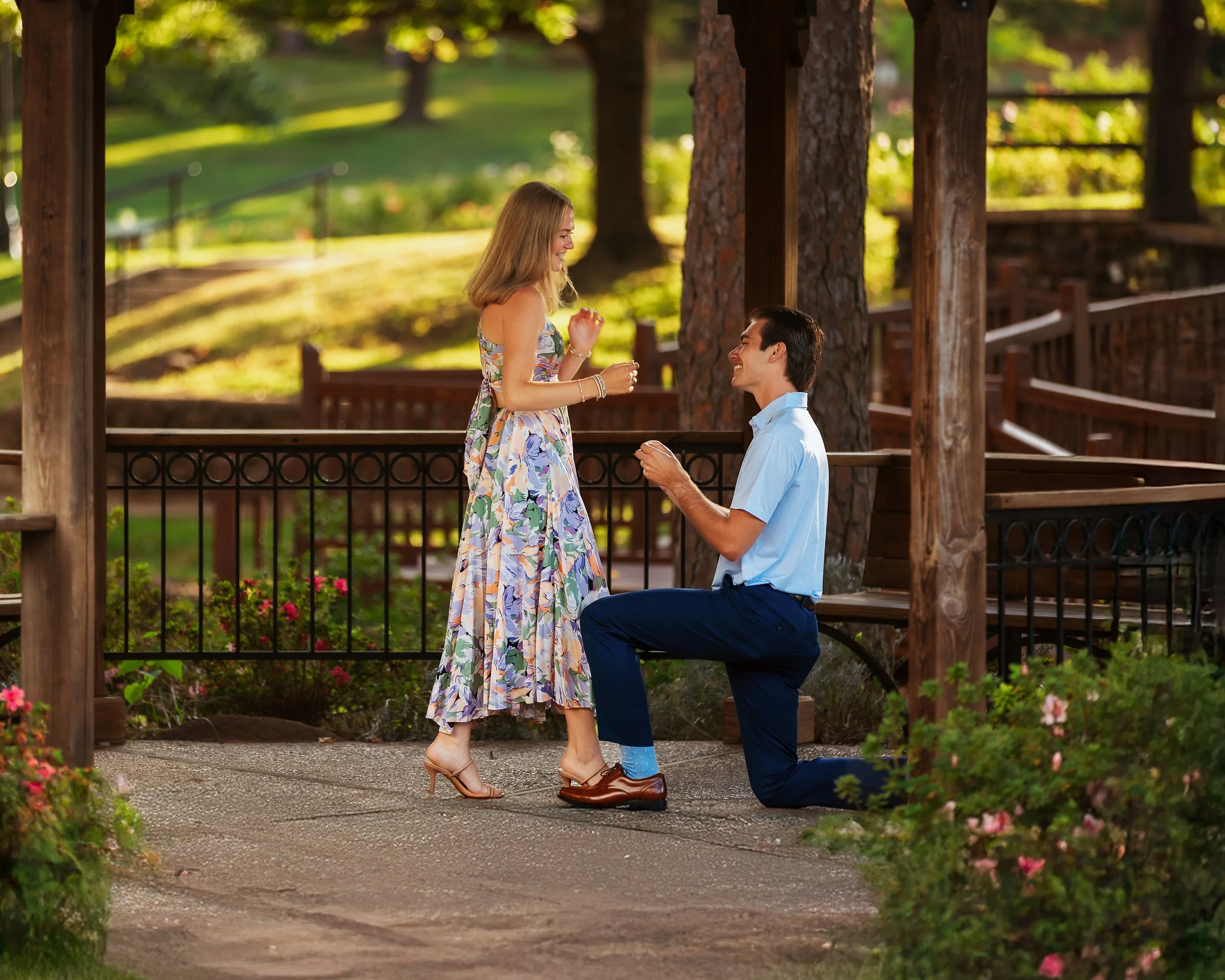 A man on one knee proposing to a woman in a park pavilion in the late afternoon, with sunlight filtering through trees and flowers in the foreground.