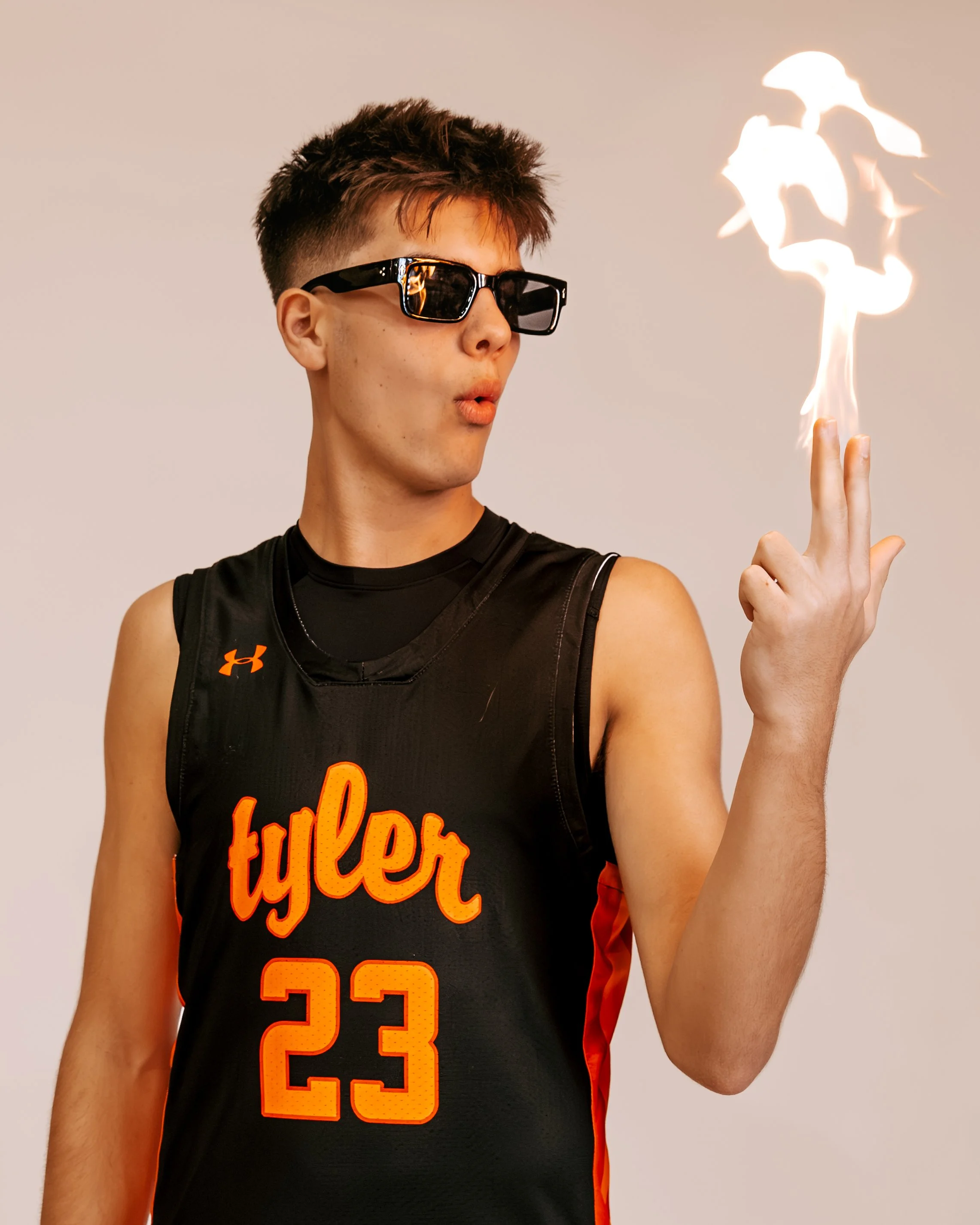 A young man in a black basketball jersey with the number 23 and the name Tyler, wearing sunglasses, is performing a fire trick with a flaming finger.