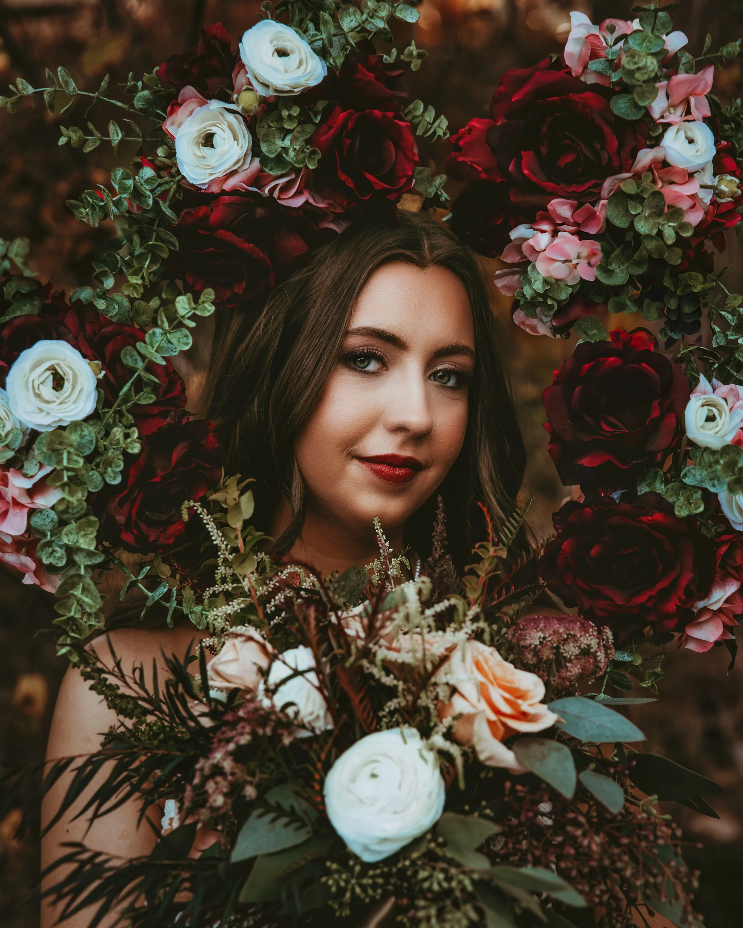 A woman with fair skin and dark hair surrounded by a large bouquet of red, white, and pink flowers, and greenery, with a blurred outdoor background.