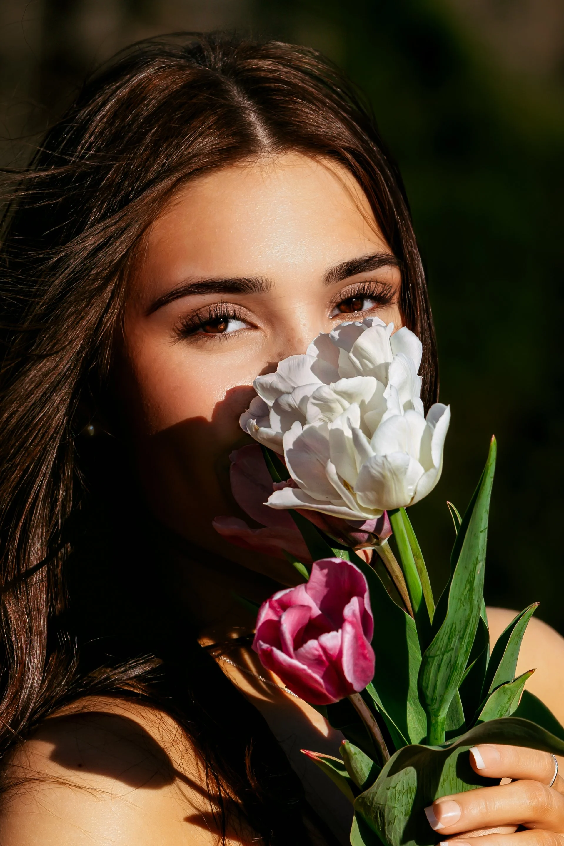 A woman with long dark hair holding a bouquet of white and pink tulips, partially covering her mouth, outdoors in sunlight.