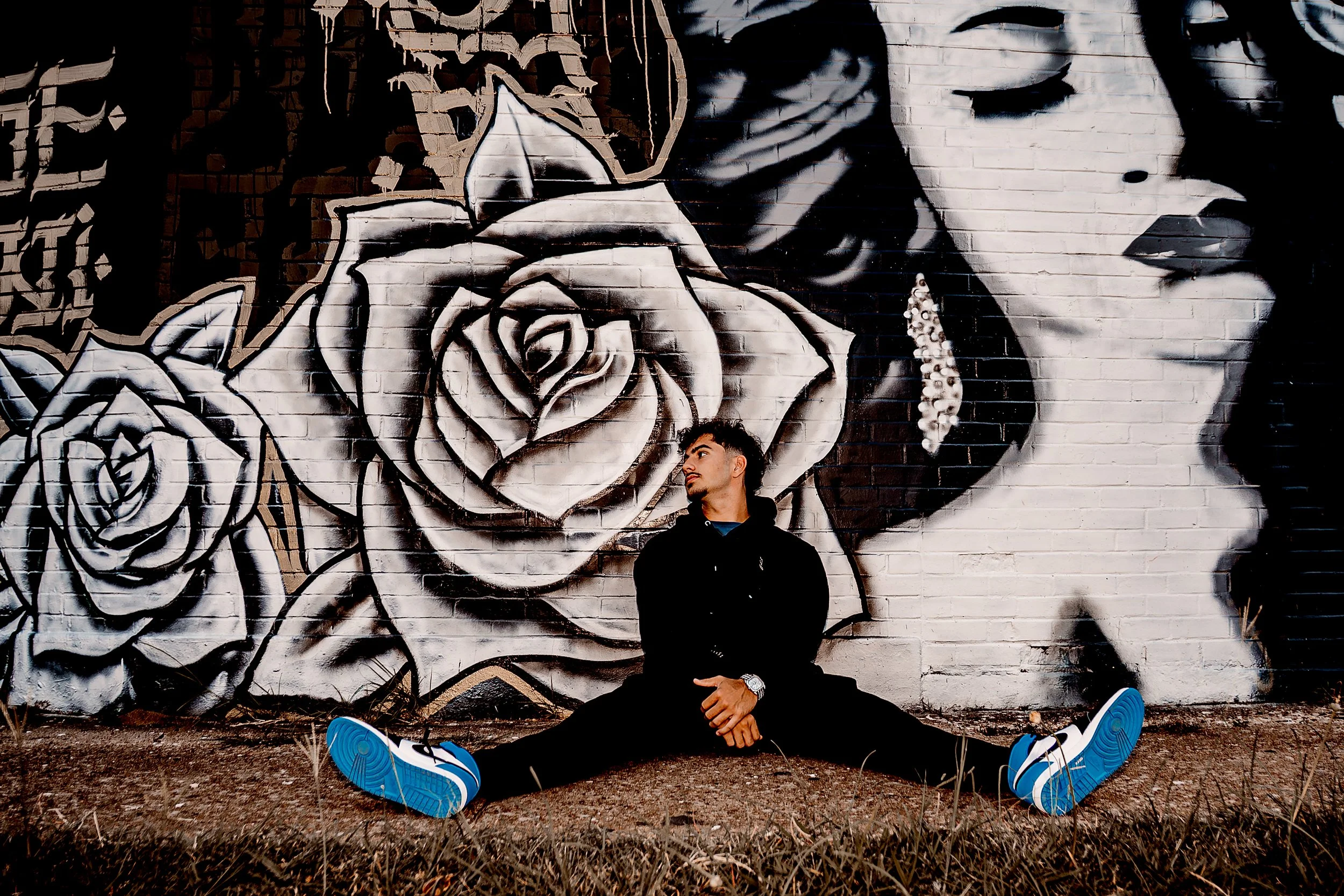 Young man sitting on the ground doing a split against a brick wall with large black and white rose portraits and a woman's face painted on it.