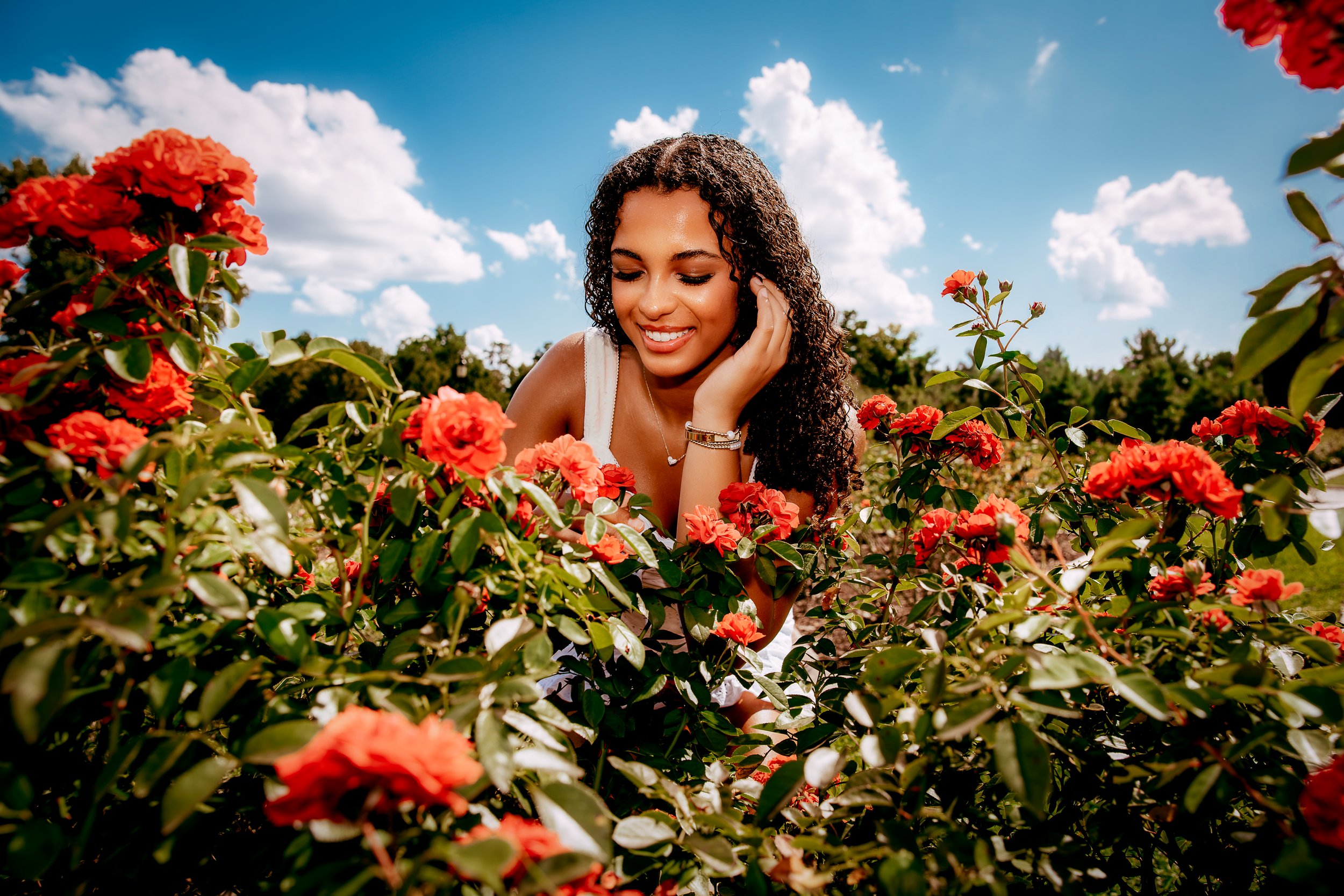 A young woman with curly hair smiling and leaning on her hand in a field of red flowers under a blue sky with fluffy clouds.