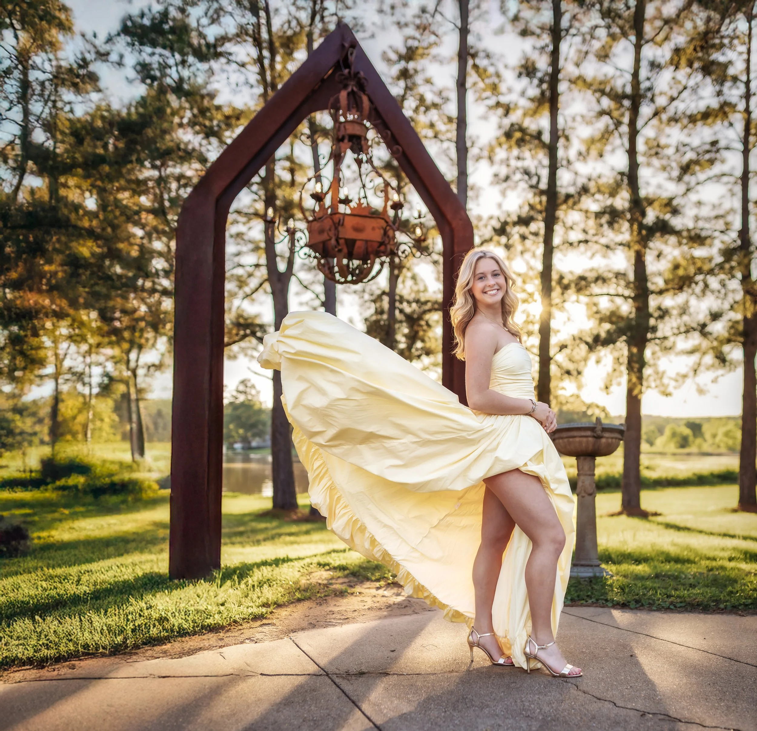 A young woman in a yellow evening gown and high heels standing on a concrete path outdoors, smiling, with a rust-colored metal archway and a chandelir hanging from it, surrounded by trees and green grass, during sunset.
