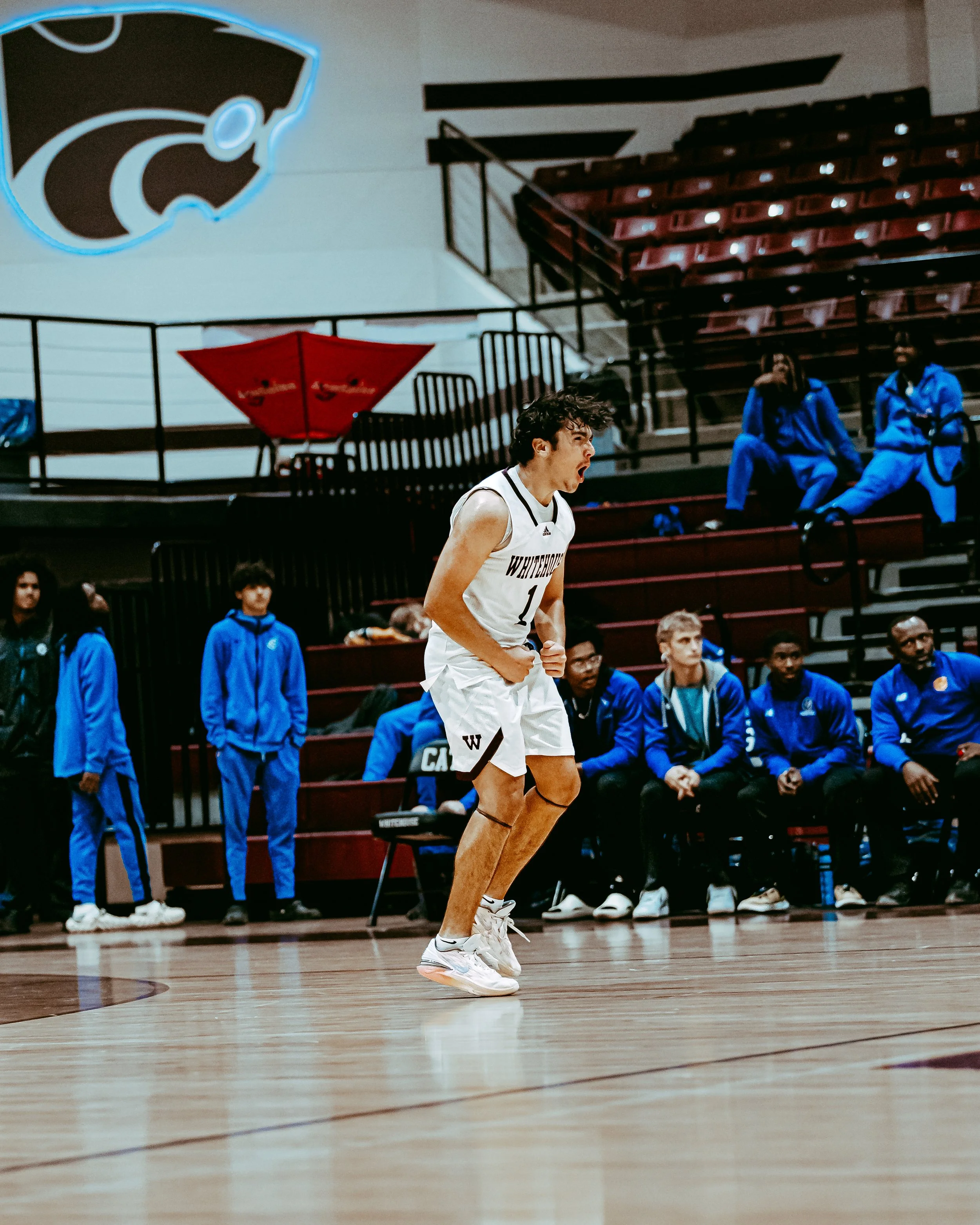 A basketball player is celebrating on the court, with teammates and coaches sitting on the bench behind him. The player is wearing a white uniform with the number 1. The gymnasium has a wooden floor and a large school logo on the wall.