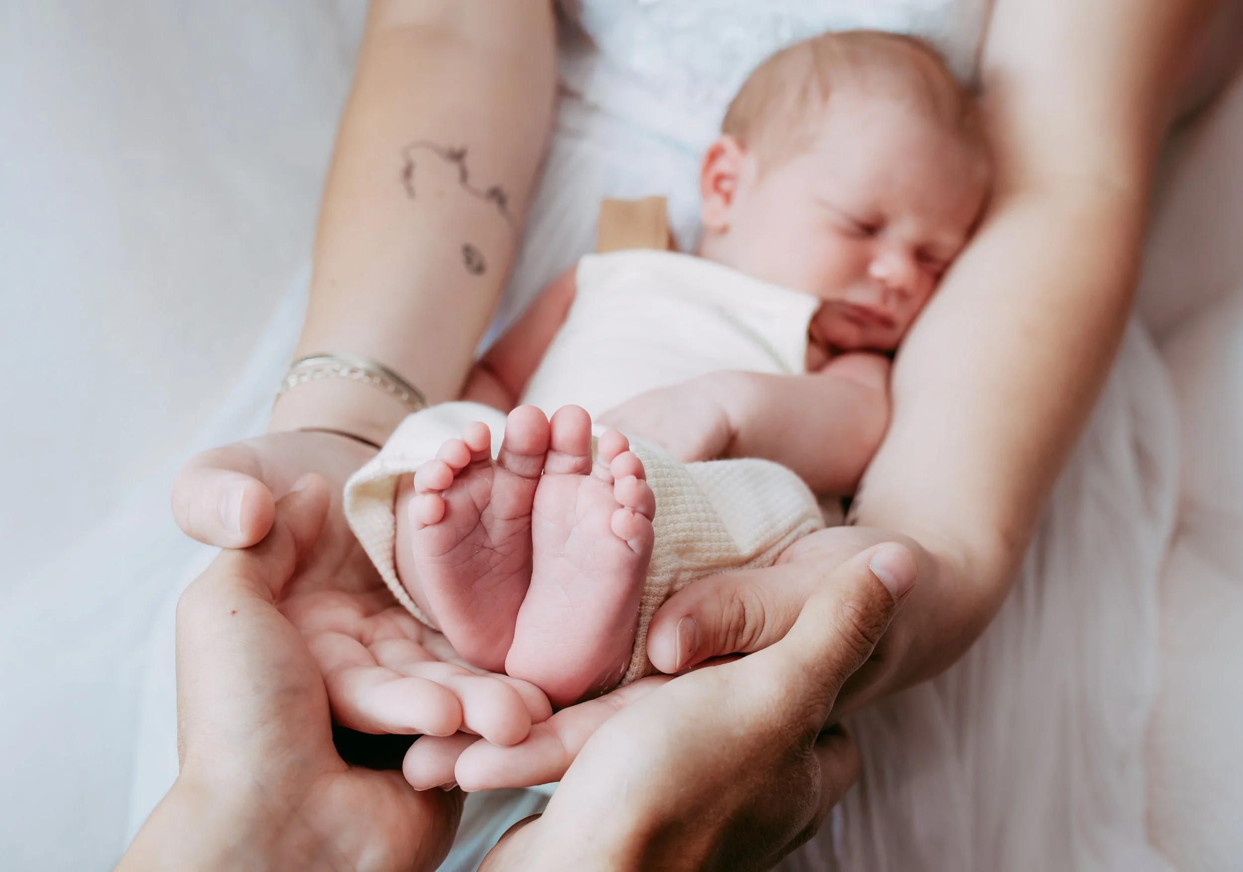 A newborn baby with reddish hair sleeping on a person's chest, with hands gently holding the baby's feet, in a tender moment.