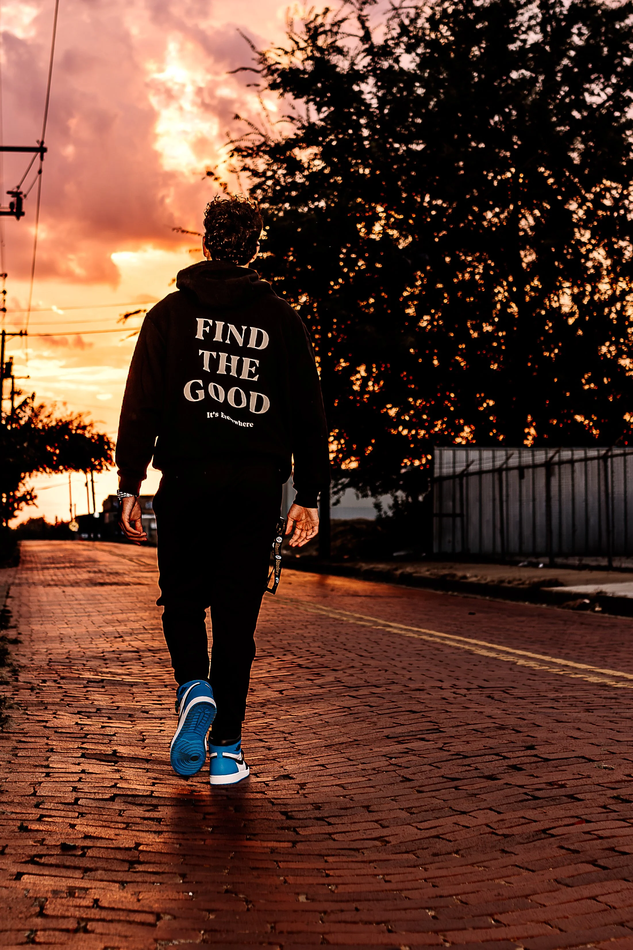 A person walking on a brick street at sunset, wearing a black hoodie with the words 'FIND THE GOOD' on the back, and bright blue sneakers.