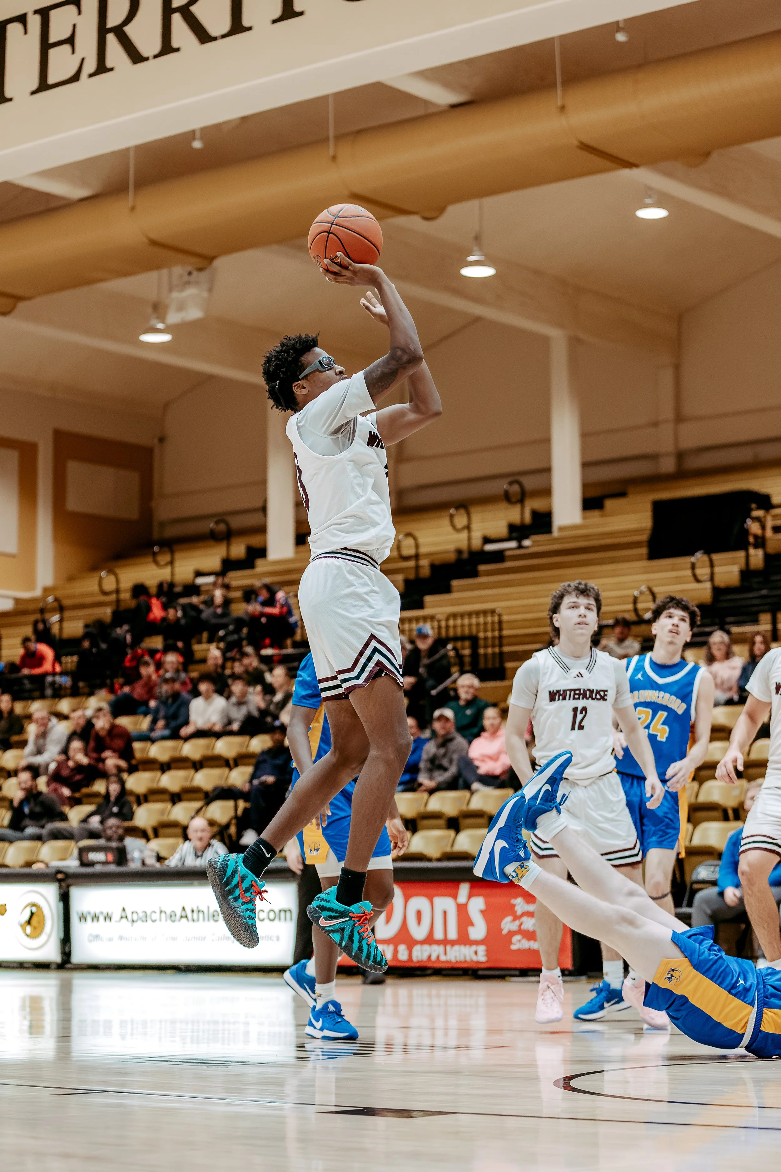 A basketball player in mid-air shooting the ball, with other players on the court and spectators in the background.
