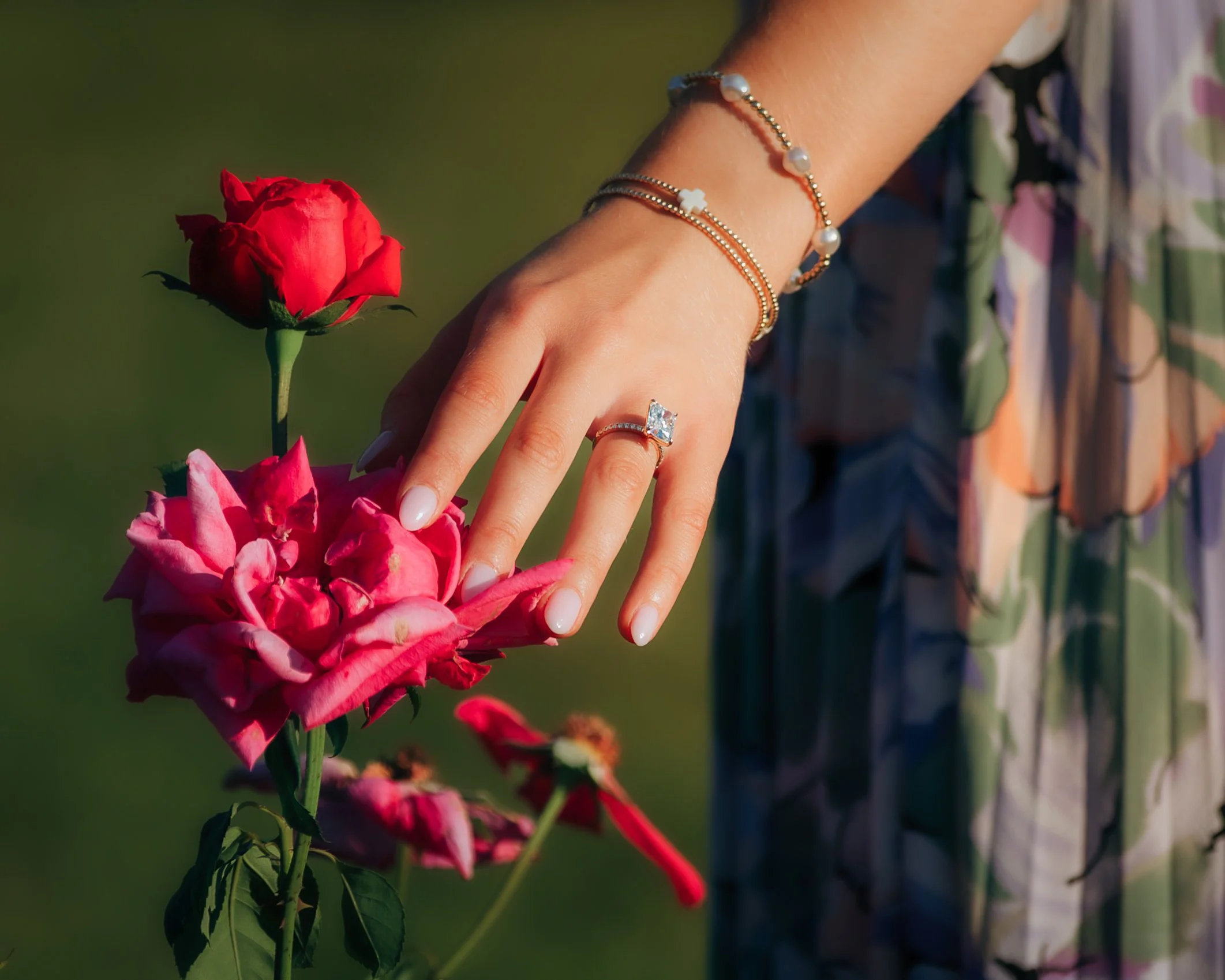 A woman's hand resting on pink and red roses, wearing a large diamond ring, with a blurred green background.