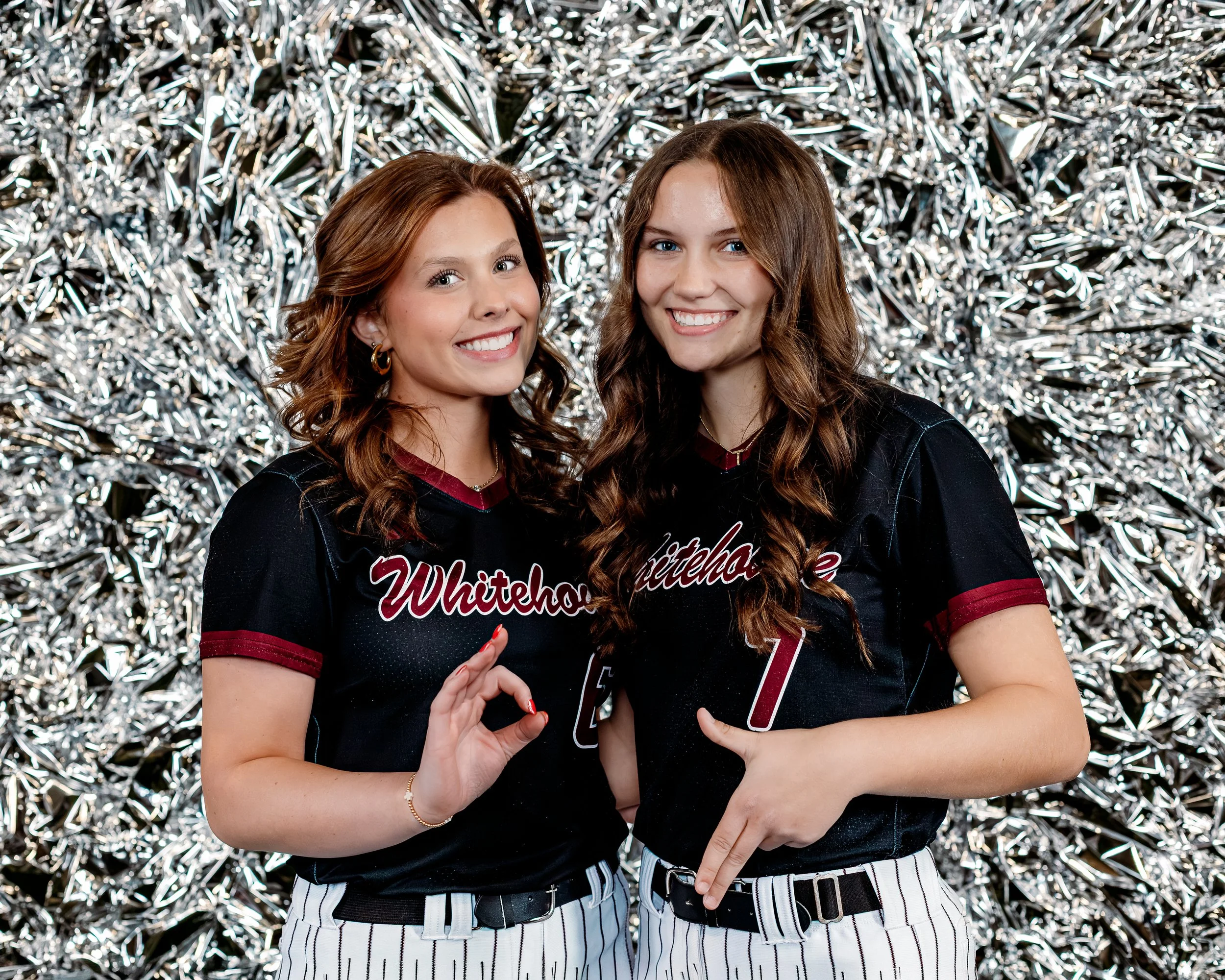 Two young women in black and white striped baseball jerseys standing in front of a shiny silver tinsel backdrop. They are smiling and pointing at the shirts which have 'Whitehorse' written on them, suggesting they are part of a team or group.